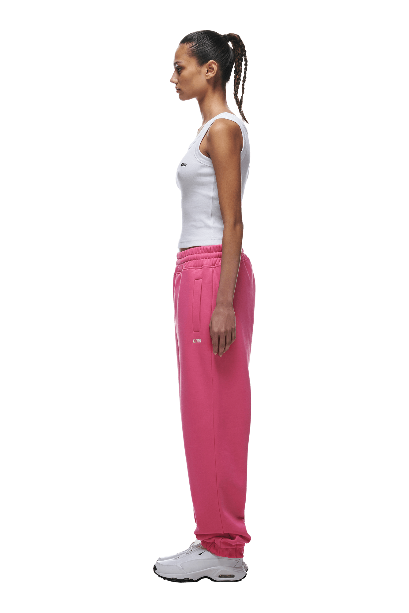 A woman with braided hair in a ponytail stands in profile, wearing a white tank top and 6PM CUFF JOGGER HOT PINK sweatpants with cuffed leg openings, paired with white sneakers, against a plain green background.