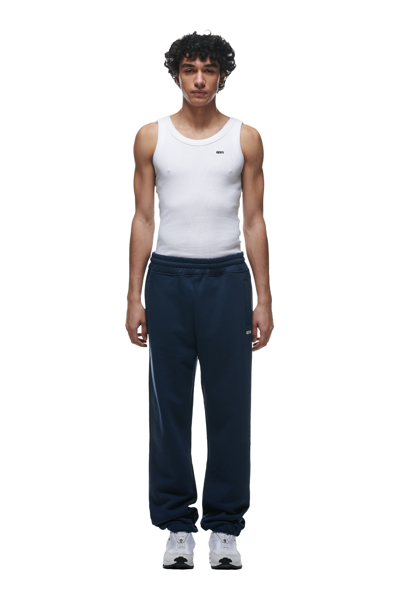 A man with short curly hair wears a white sleeveless ribbed tank, 6PM CUFF JOGGER NAVY sweatpants with cuffed hems, and white sneakers. He stands facing forward against a plain background, arms at his sides.