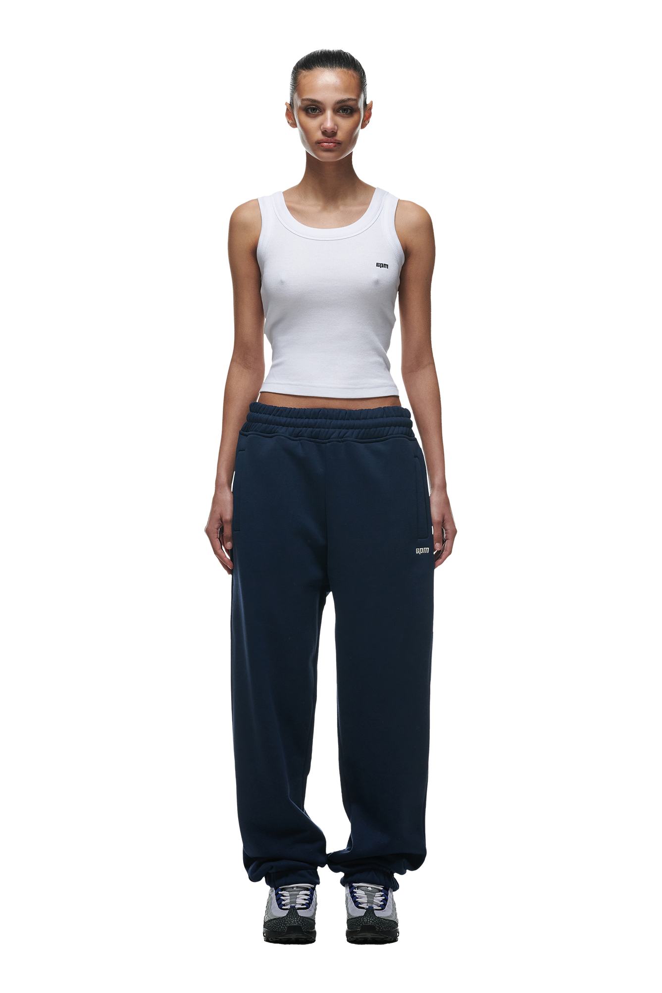 A woman stands facing forward wearing a white tank top, 6PM CUFF JOGGER NAVY sweatpants with cuffed leg openings, and black sneakers. Her hair is pulled back and the background is plain white.