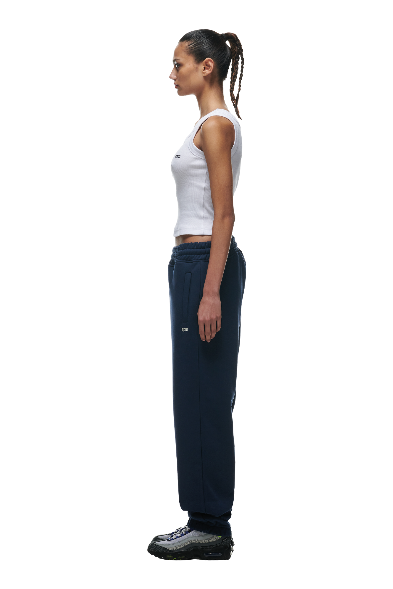 A woman with her hair in a ponytail stands in profile against a white background, wearing a white sleeveless tank top, black sneakers, and the 6PM CUFF JOGGER NAVY sweatpants with classic fit and cuffed leg opening.