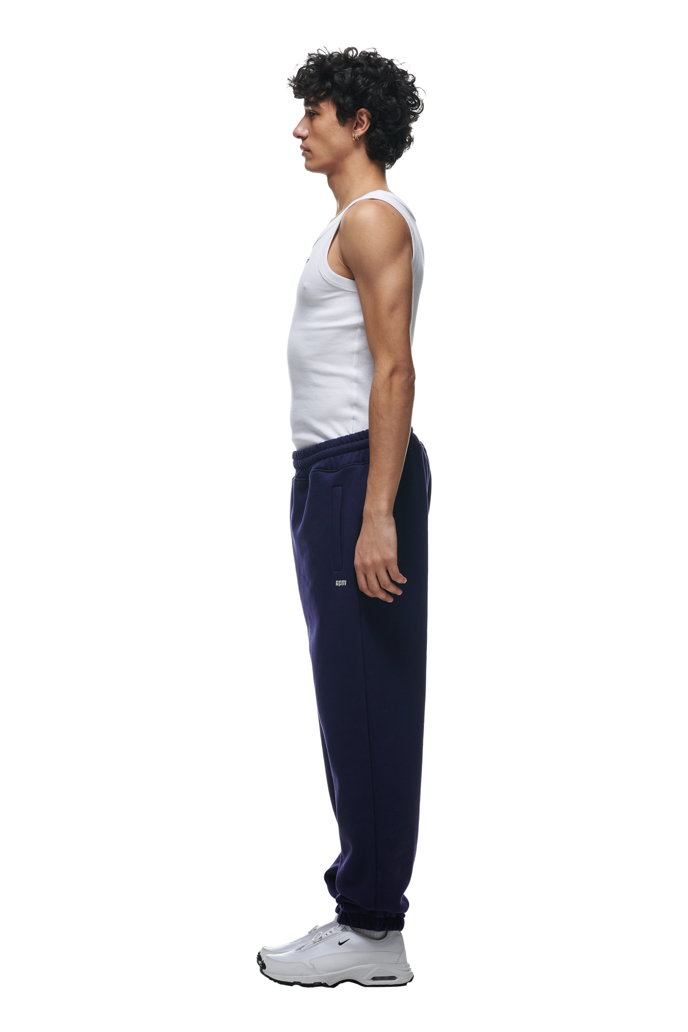 A person stands in profile, facing left, wearing 6PM CUFF JOGGER ORBIT BLUE sweatpants with a classic fit and cuffed leg opening, paired with a white tank top and white sneakers against a plain white background.