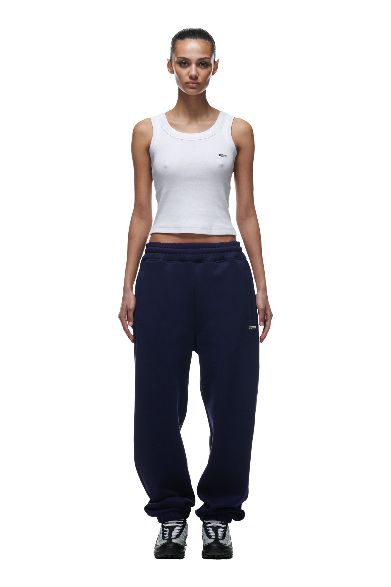 A woman with a neutral expression and tied-back hair faces forward, wearing a white sleeveless tank top, 6PM CUFF JOGGER ORBIT BLUE sweatpants with cuffed legs, and black and white sneakers.