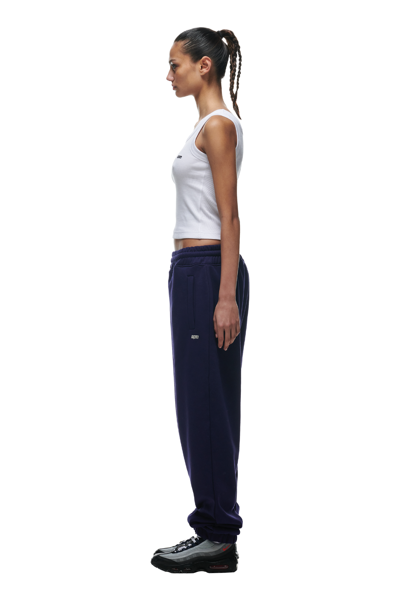 A woman with cornrow braids stands in profile wearing a white tank top, 6PM CUFF JOGGER ORBIT BLUE sweatpants with cuffed legs, and black sneakers. She looks ahead with her arms relaxed at her sides.