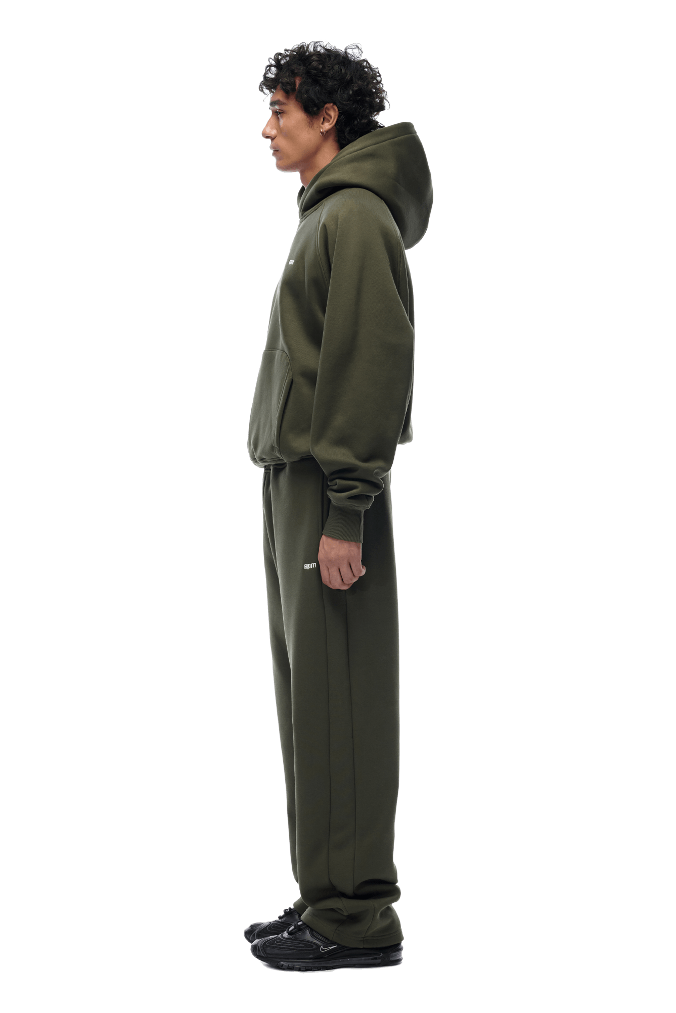 A person stands in profile, facing left, wearing the 6PM HOODIE FOREST NIGHT and matching sweatpants with black sneakers. The hood is up for everyday use. The background is transparent.