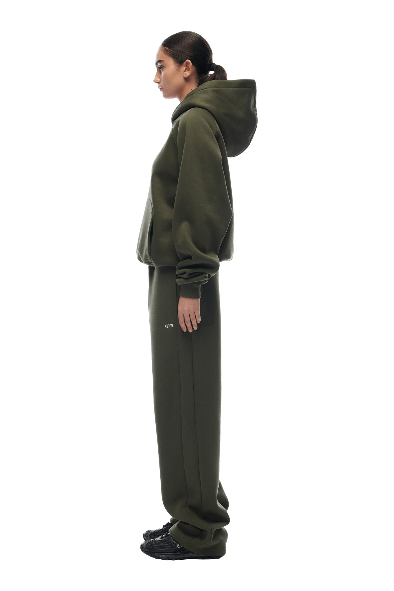 A person stands in profile wearing the 6PM HOODIE FOREST NIGHT, an oversized dark green raglan-sleeved hoodie with matching sweatpants and black sneakers. The outfit is casual and comfortable, suitable for everyday wear. Transparent background.