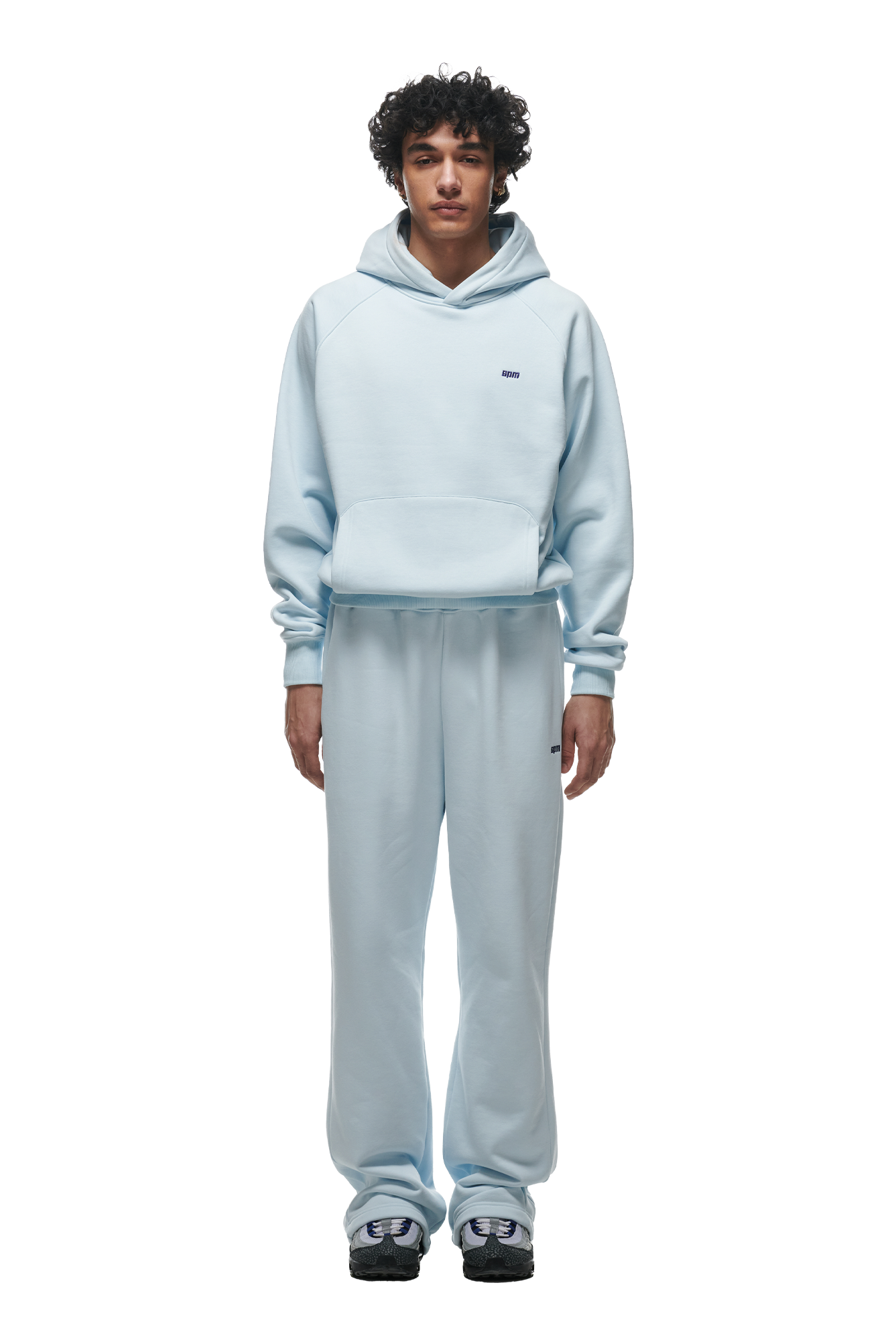 A person stands facing forward against a white background, wearing the 6PM HOODIE ICE BLUE with raglan sleeves, kangaroo pocket, matching sweatpants, and white-black sneakers—a casual outfit ideal for everyday wear.