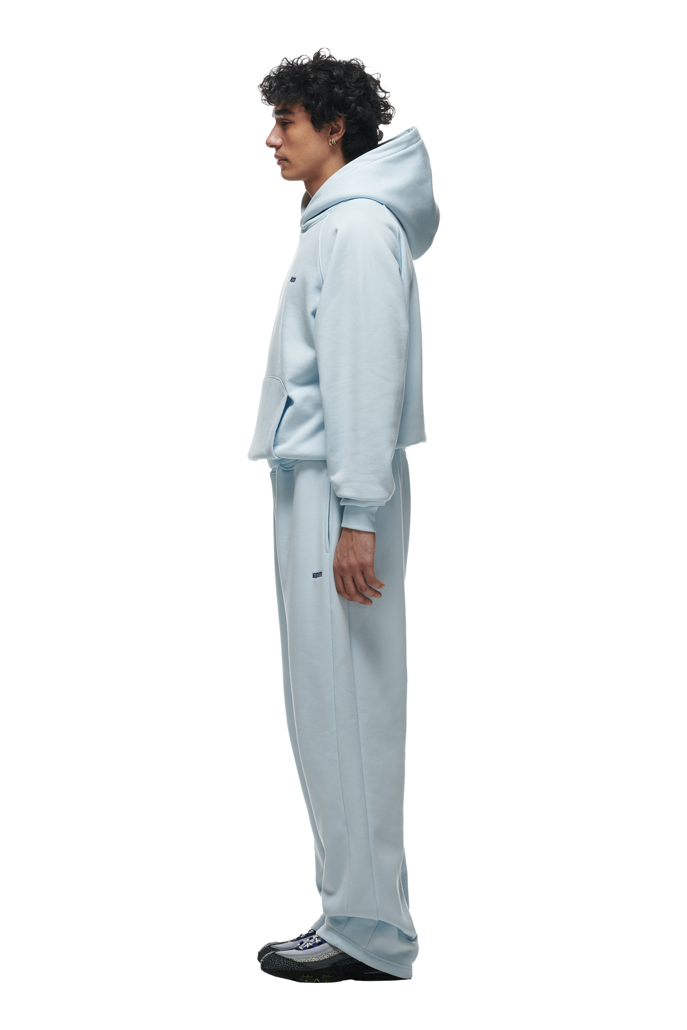 A person in profile wears the 6PM HOODIE ICE BLUE with Raglan sleeves, a kangaroo pocket, and matching sweatpants, hands relaxed at their sides and black shoes—an everyday look set against a plain white background.