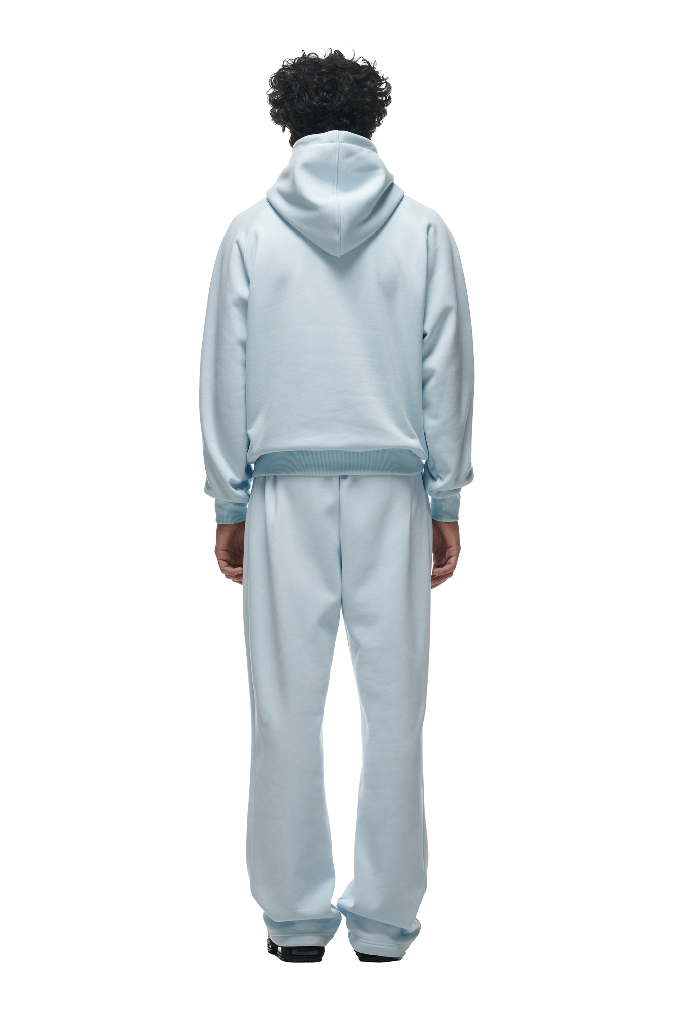 A person with curly hair is seen from behind wearing the 6PM HOODIE ICE BLUE, featuring Raglan sleeves, a kangaroo pocket, and matching sweatpants with black shoes against a white background—ideal for daily wear.