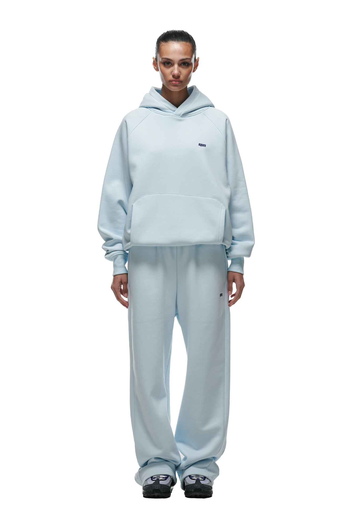 A woman faces forward in the 6PM HOODIE ICE BLUE—featuring raglan sleeves, kangaroo pocket, and matching wide-leg sweatpants. She pairs the relaxed set with black sneakers; her hair is tied back, expression neutral. Perfect for everyday wear.