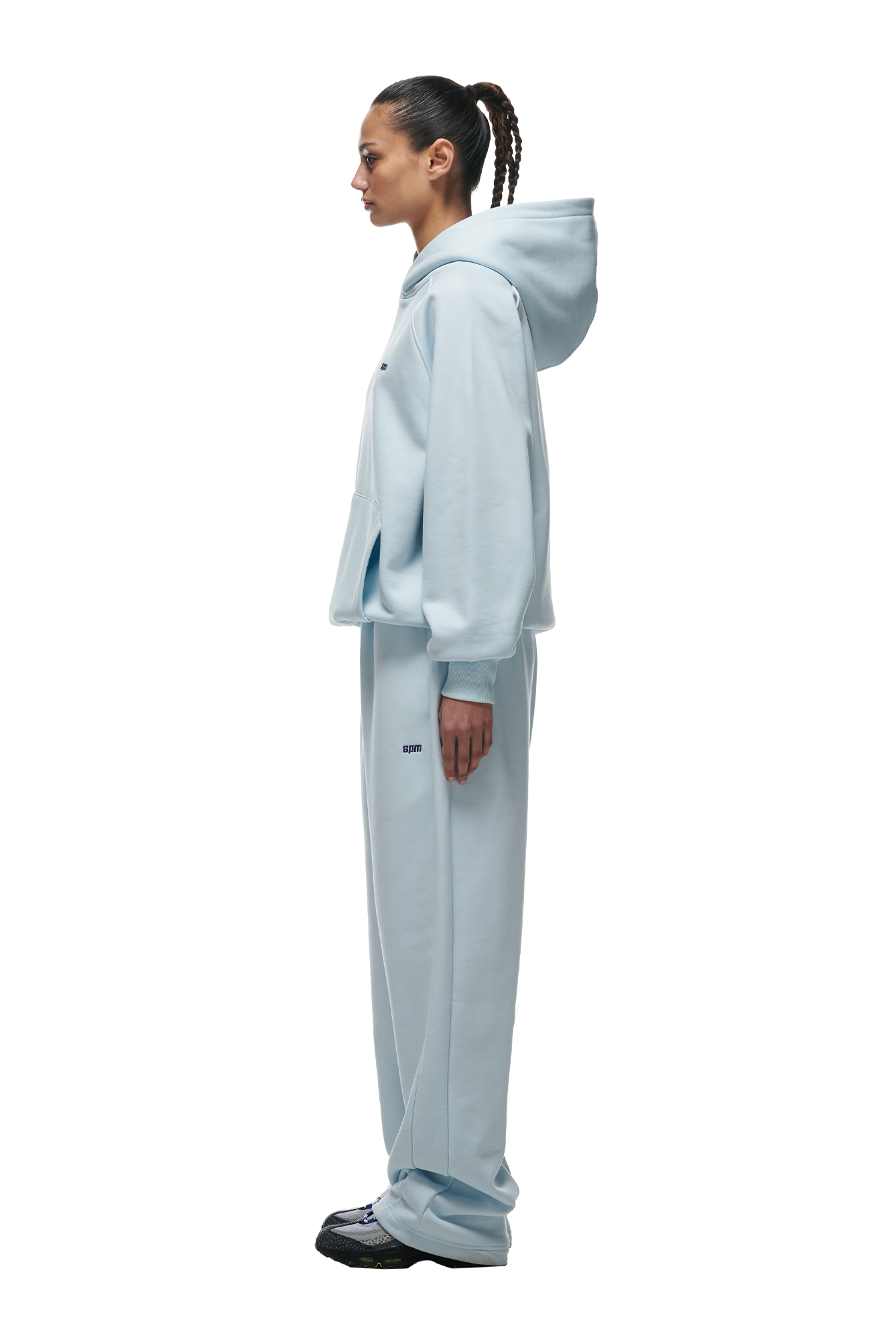 A woman in profile models the 6PM HOODIE ICE BLUE—a light blue raglan-sleeved hoodie with a kangaroo pocket, paired with matching sweatpants and black sneakers—against a white background.