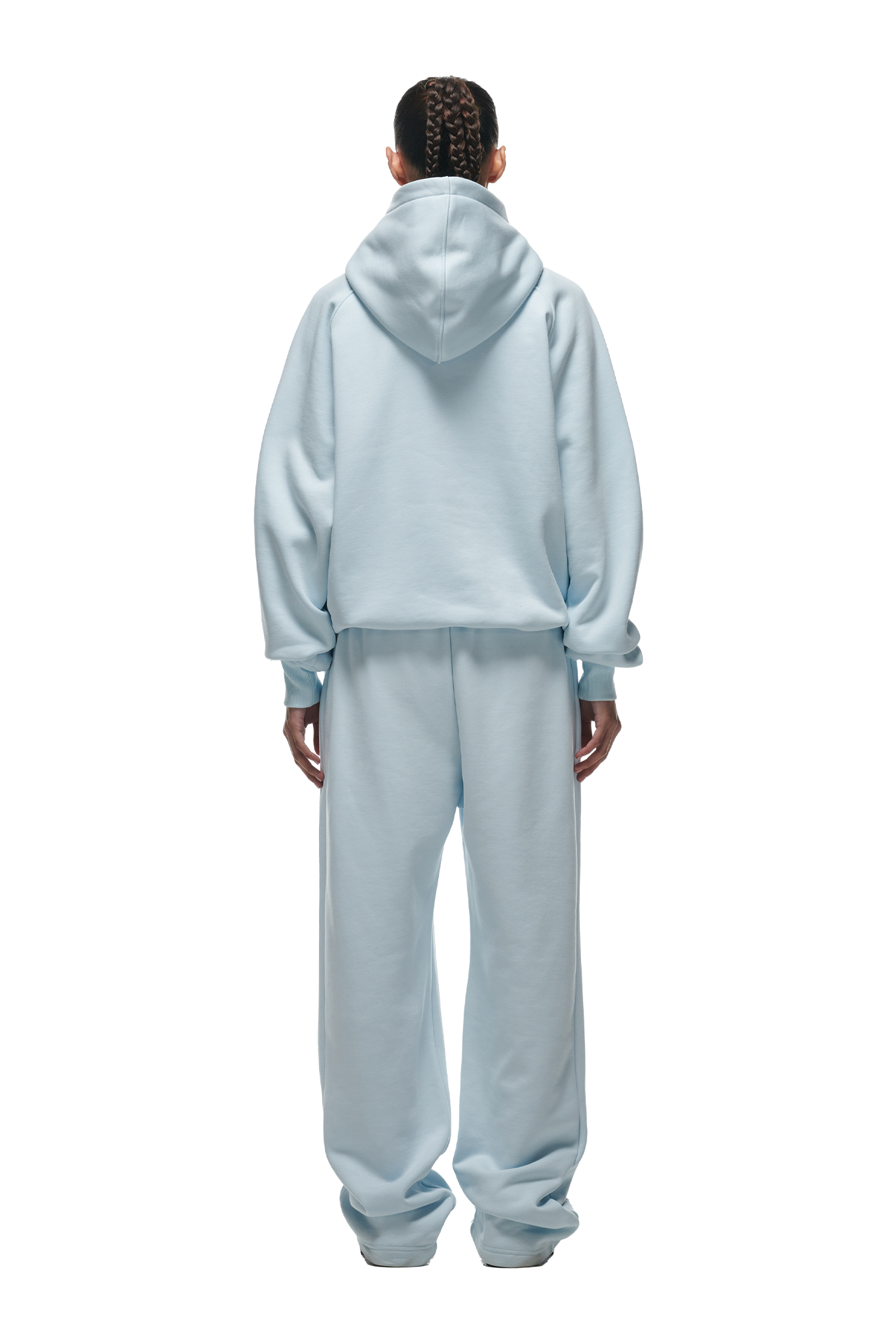 Person with braided hair stands facing away, wearing 6PM’s HOODIE ICE BLUE—an ice blue, loose-fit raglan hoodie paired with matching sweatpants. The outfit offers a relaxed fit ideal for everyday wear, shown against a white background.