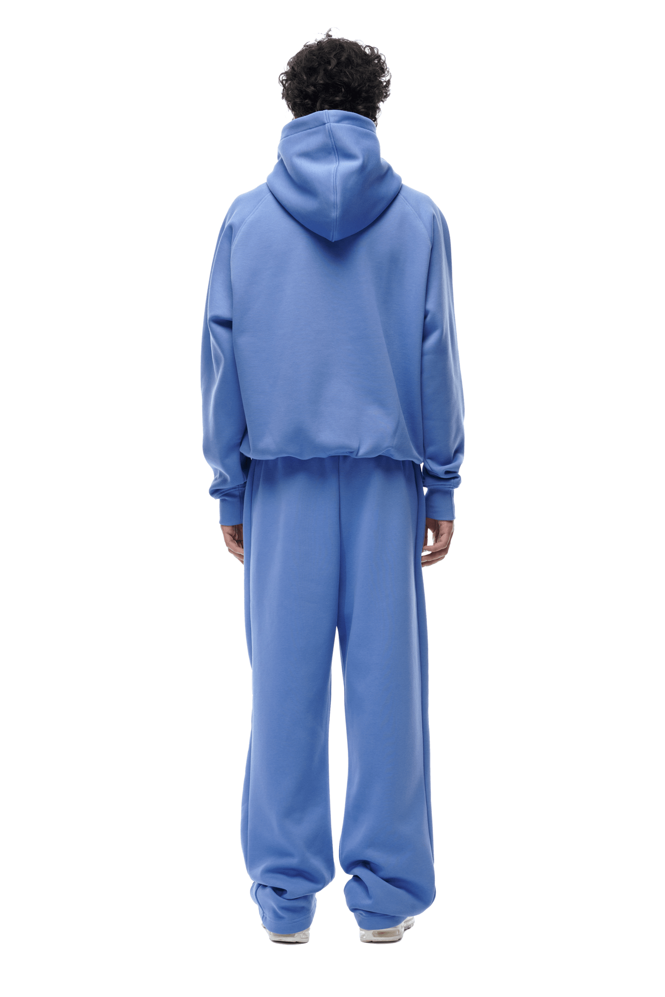 A person with short curly hair faces away, wearing the 6PM HOODIE PERSIAN BLUE with raglan sleeves and a kangaroo pocket, paired with matching sweatpants and white sneakers. The transparent background suits everyday use.