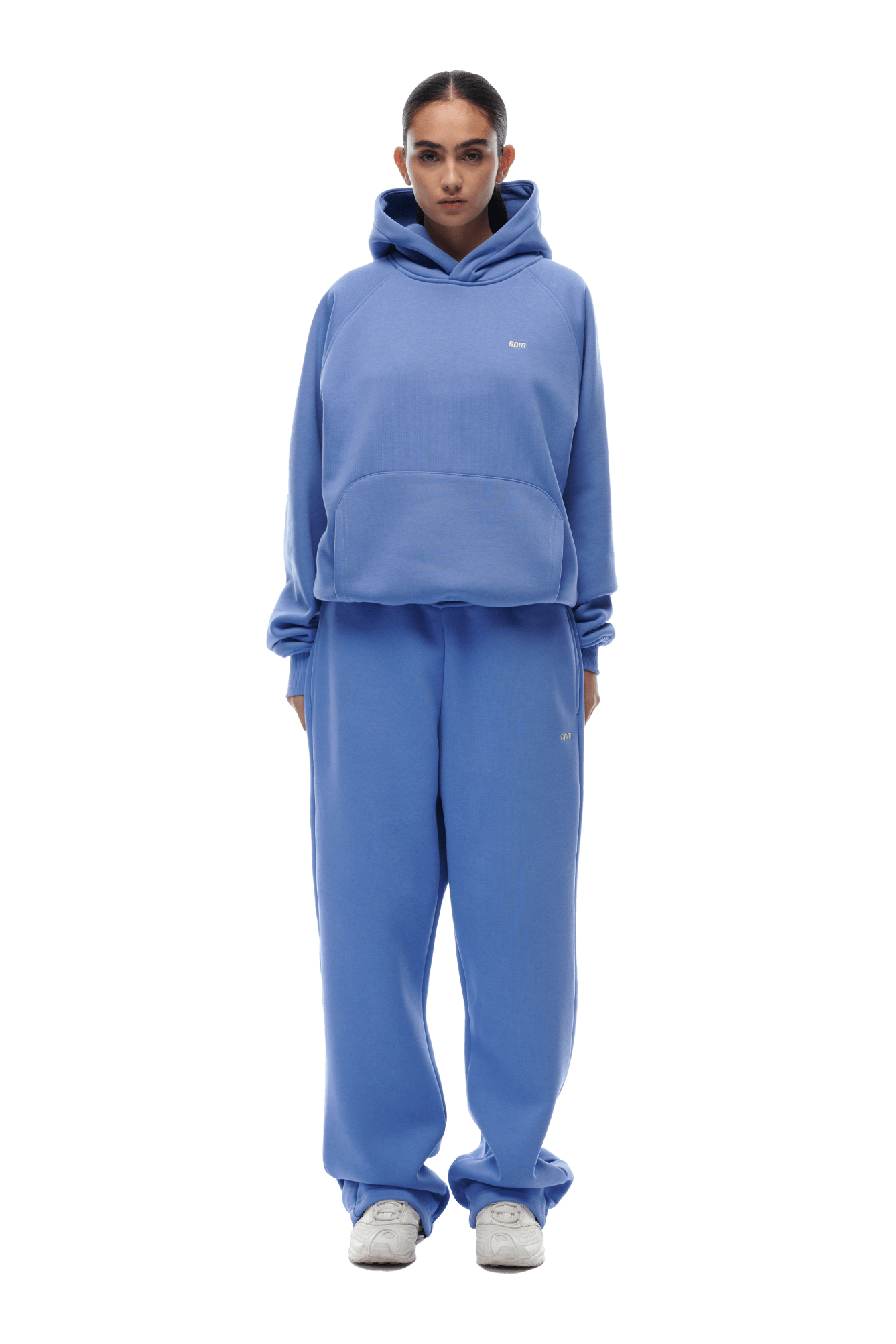 A person stands facing forward, wearing the 6PM HOODIE PERSIAN BLUE—a loose-fitting raglan-sleeve hoodie with kangaroo pocket and matching sweatpants. Hair pulled back, hands at sides, white sneakers. Transparent background suitable for everyday use.