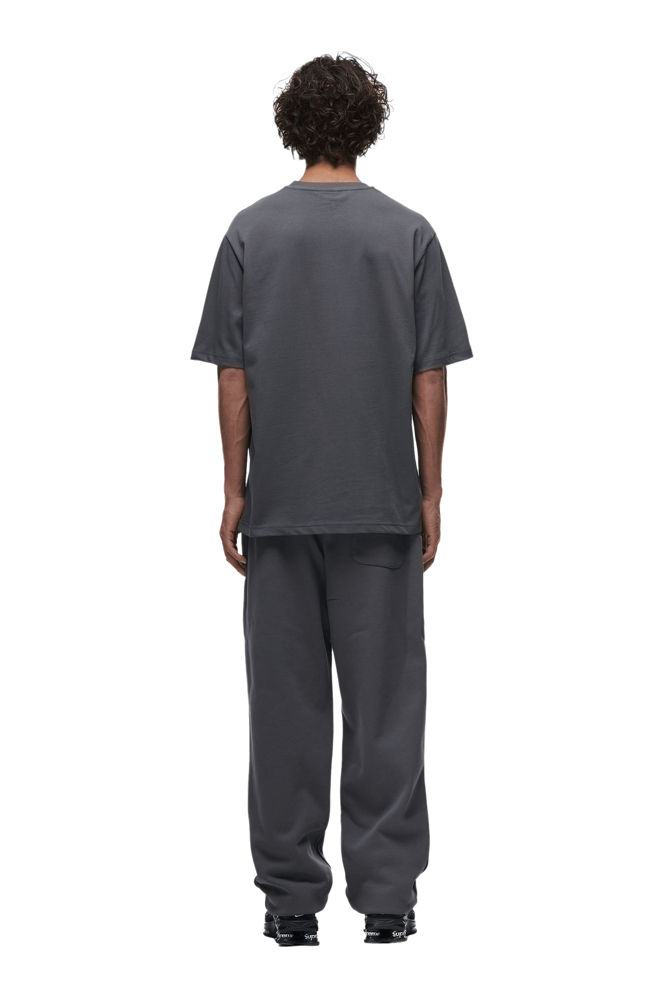 A person with short curly hair faces away, wearing the 6PM T-SHIRT ANTHRACITE and matching sweatpants with black sneakers, set against a plain green background.