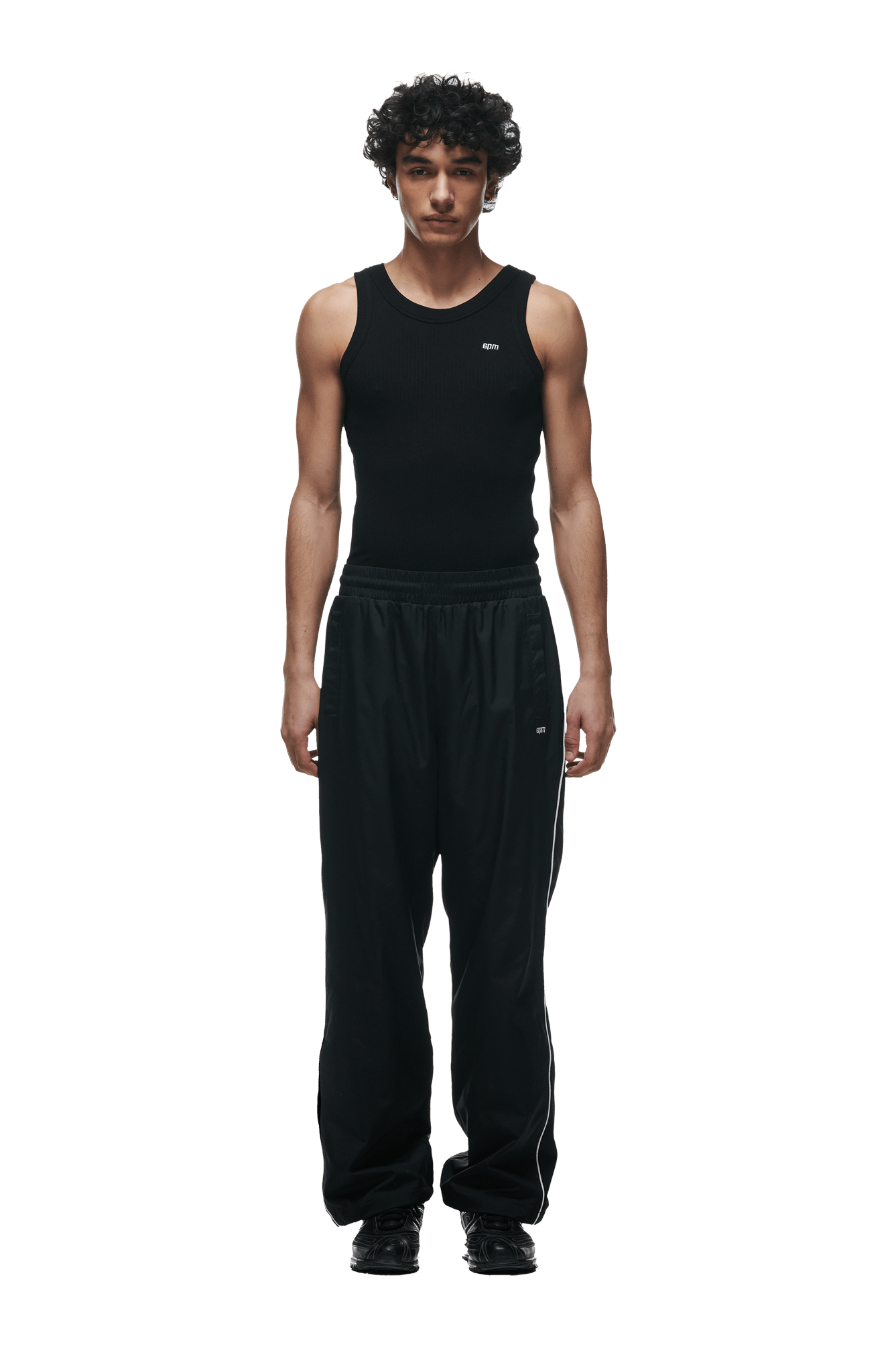 A young man with curly hair faces forward, wearing a black sleeveless tank top, 6PM 6PM+ NYLON PANTS OPEN LEG BLACK with zipped pockets and side piping, and black shoes, against a plain green background.