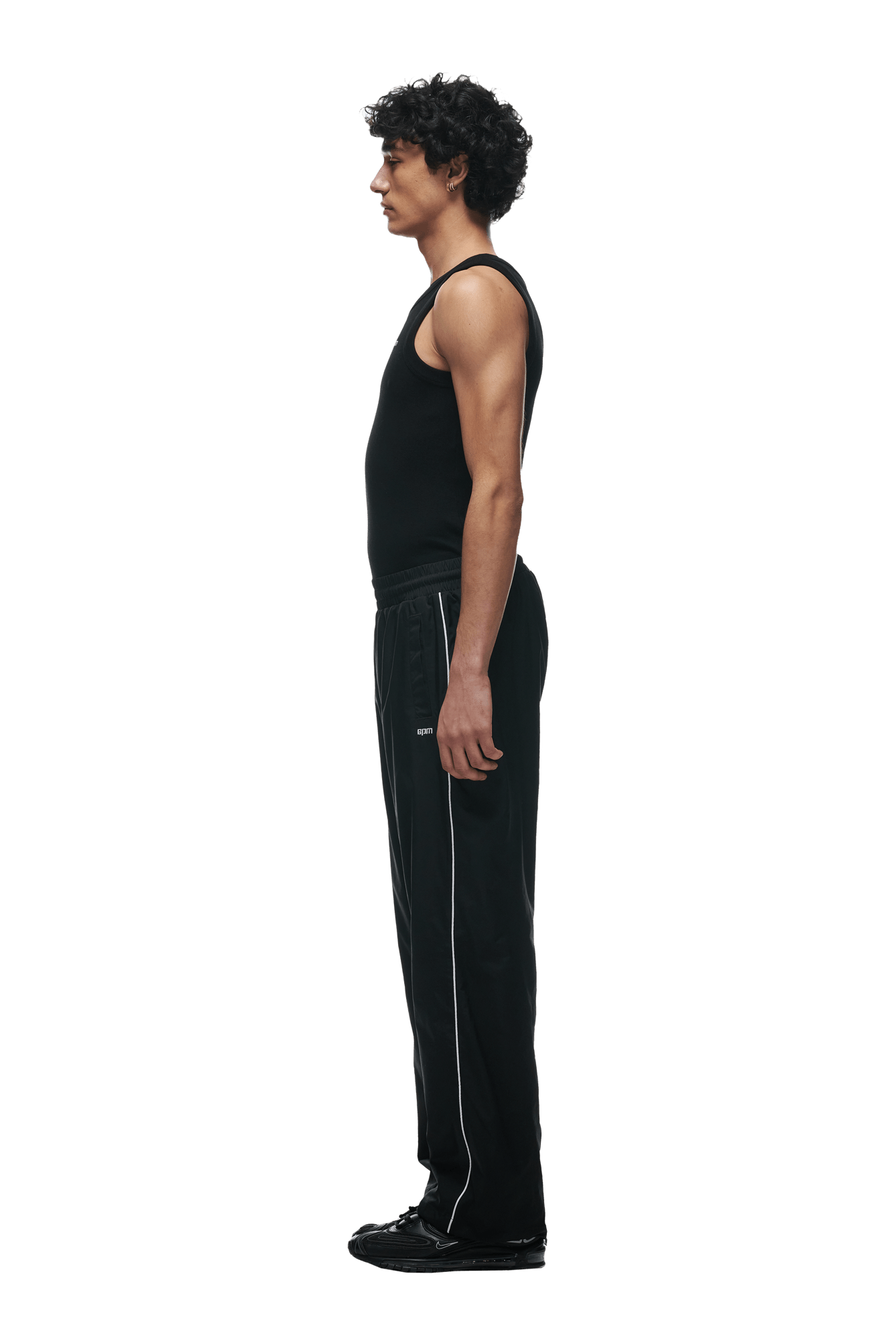 A person with short curly hair stands in profile facing left, wearing a black sleeveless shirt, 6PM 6PM+ NYLON PANTS OPEN LEG BLACK with white side stripes and zipped pockets, and black shoes, on a plain green background.