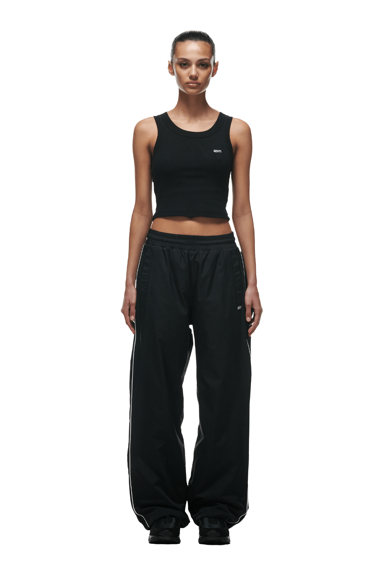 A woman faces forward, wearing a black sleeveless crop top and 6PM+ NYLON PANTS OPEN LEG BLACK by 6PM, featuring subtle side stripes and zipped pockets. Her hair is tied back, she wears black shoes, and stands against a plain green background.
