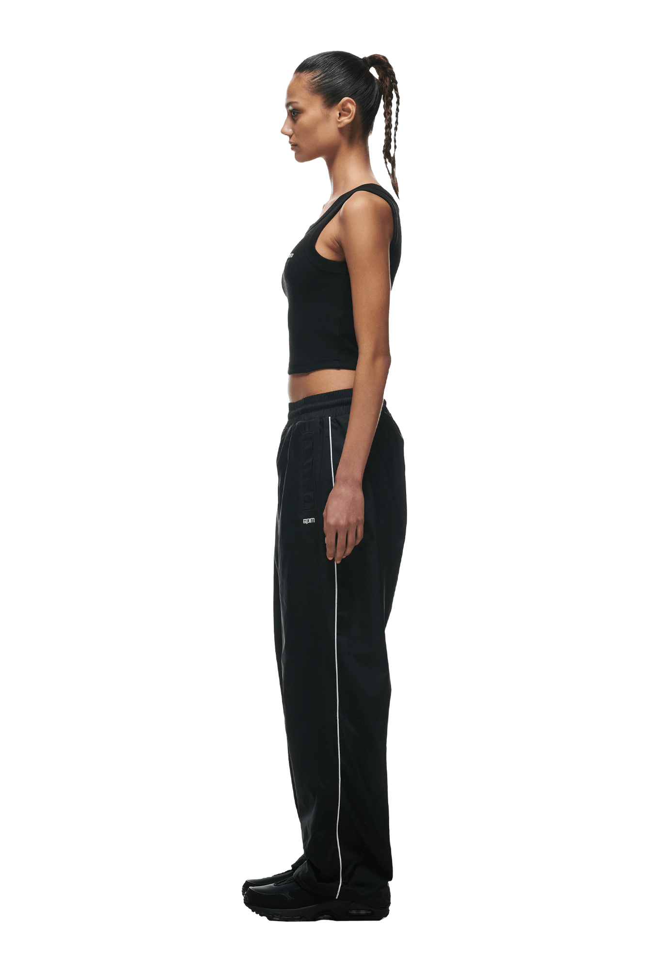 A woman with a braided ponytail stands in profile, wearing a black tank top and 6PM+ Nylon Pants Open Leg Black by 6PM featuring white piping, zipped pockets, and an adjustable drawstring, along with black shoes, against a transparent background.