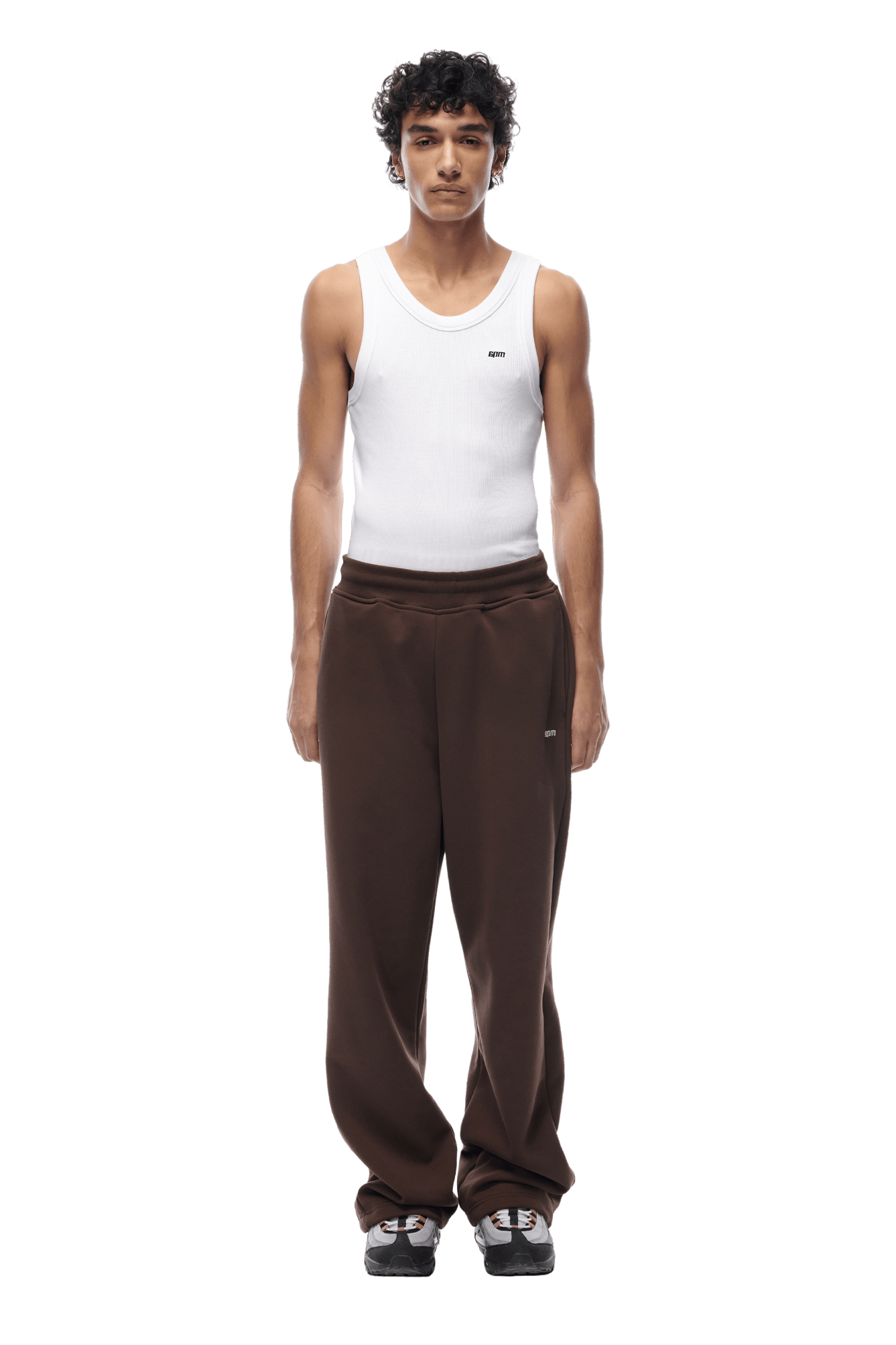 A person with short curly hair wears a white sleeveless tank top, OPEN LEG JOGGER CHOCOLATE BROWN pants with stacking length, and black sneakers, standing facing forward against a transparent background.