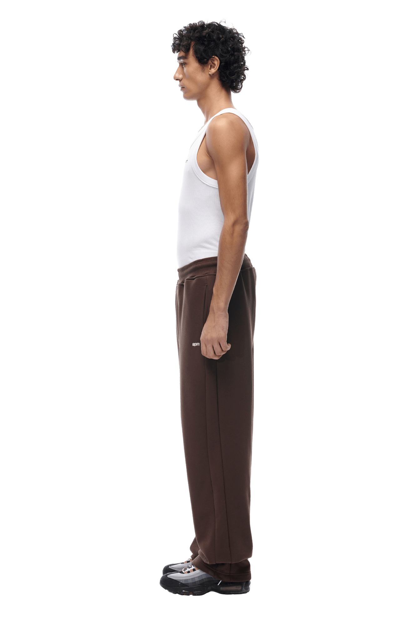 A man with curly hair stands in profile, wearing a white tank top, black sneakers, and the OPEN LEG JOGGER CHOCOLATE BROWN pants against a plain background.