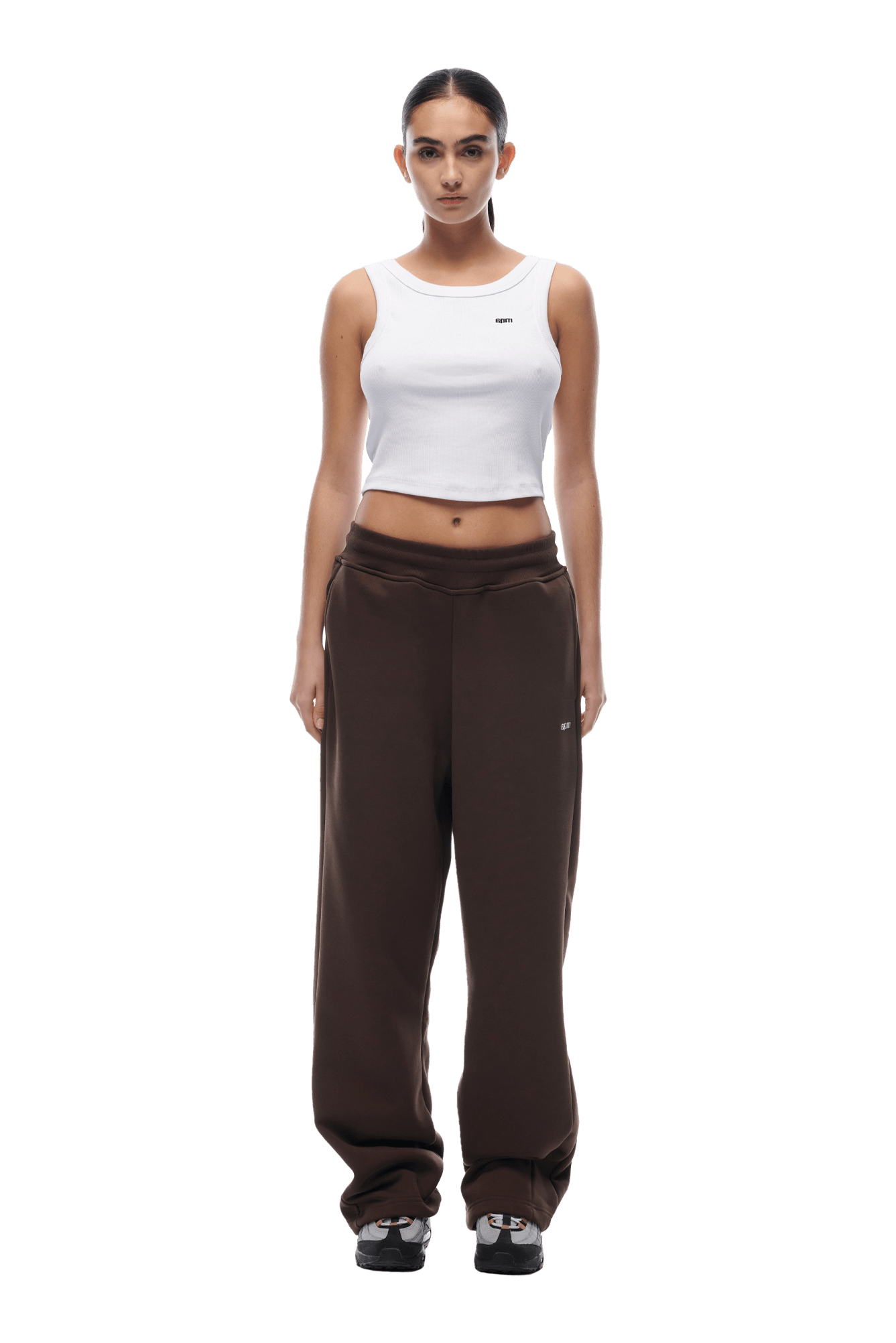 A woman faces forward in a white sleeveless crop top and Open Leg Jogger Chocolate Brown pants with stacking length, paired with black sneakers. Her dark hair is tied back, and she poses against a plain background with a neutral expression.