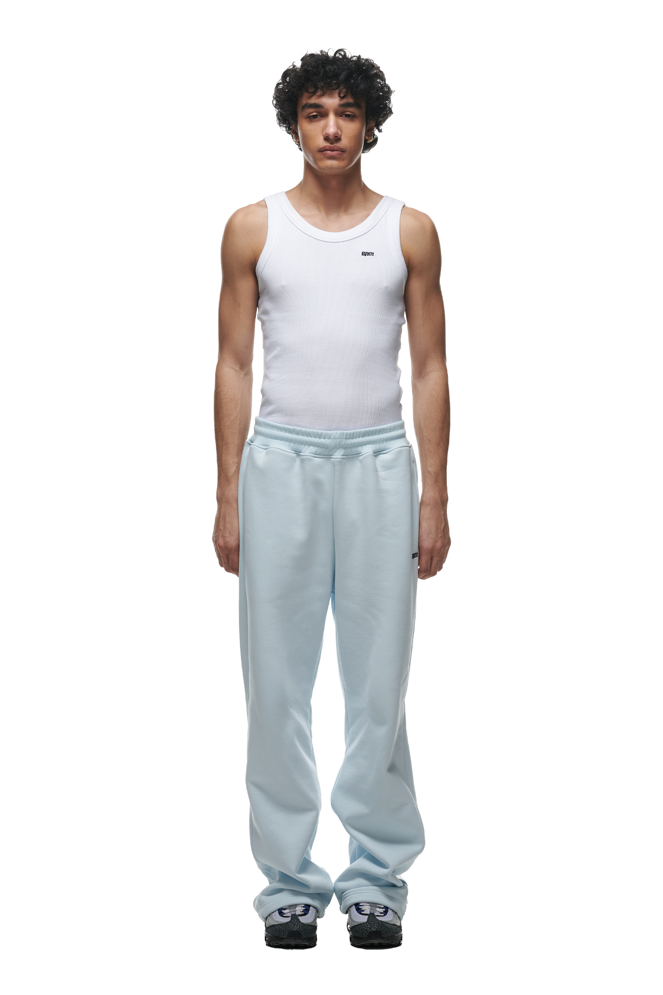 A man with curly hair faces the camera against a white background, wearing 6PMs OPEN LEG JOGGER ICE BLUE, a white sleeveless ribbed tank top, and black and white sneakers.