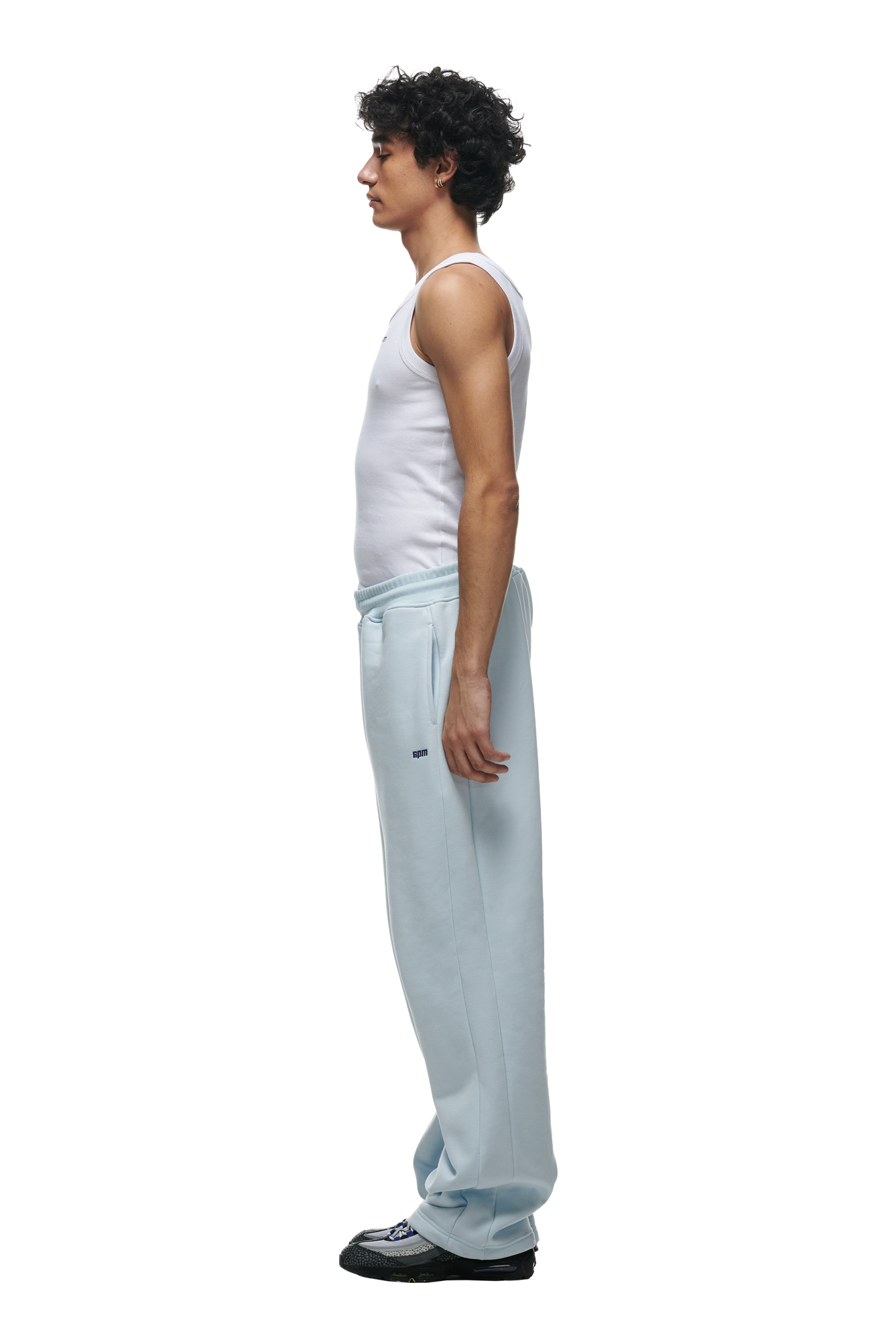 A man with curly hair stands in profile, wearing a white tank top and 6PM OPEN LEG JOGGER ICE BLUE sweatpants with stacking length, paired with black shoes, creating a modern classic look against a white background.
