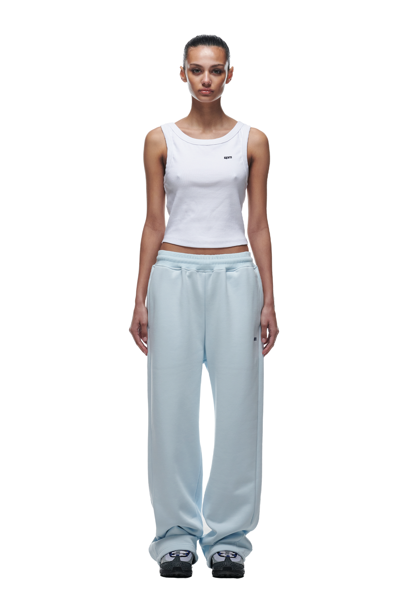 A woman with her hair tied back and a neutral expression wears the 6PM OPEN LEG JOGGER ICE BLUE sweatpants, styled with a white sleeveless tank top and black-and-white sneakers, standing facing forward.