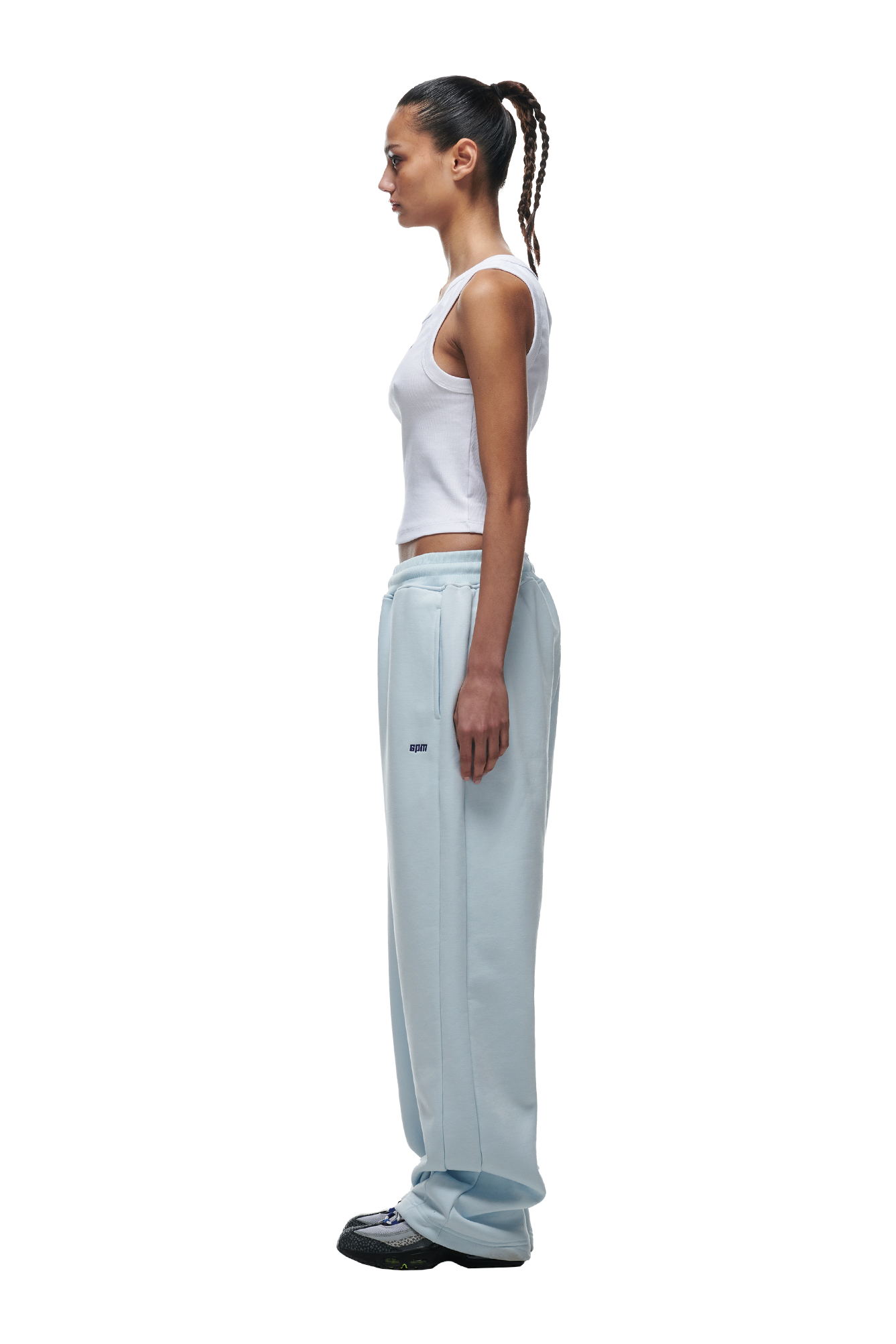 A woman with a braided ponytail stands in profile, wearing a white tank top, 6PM OPEN LEG JOGGER ICE BLUE pants, and black-and-white sneakers against a white background.