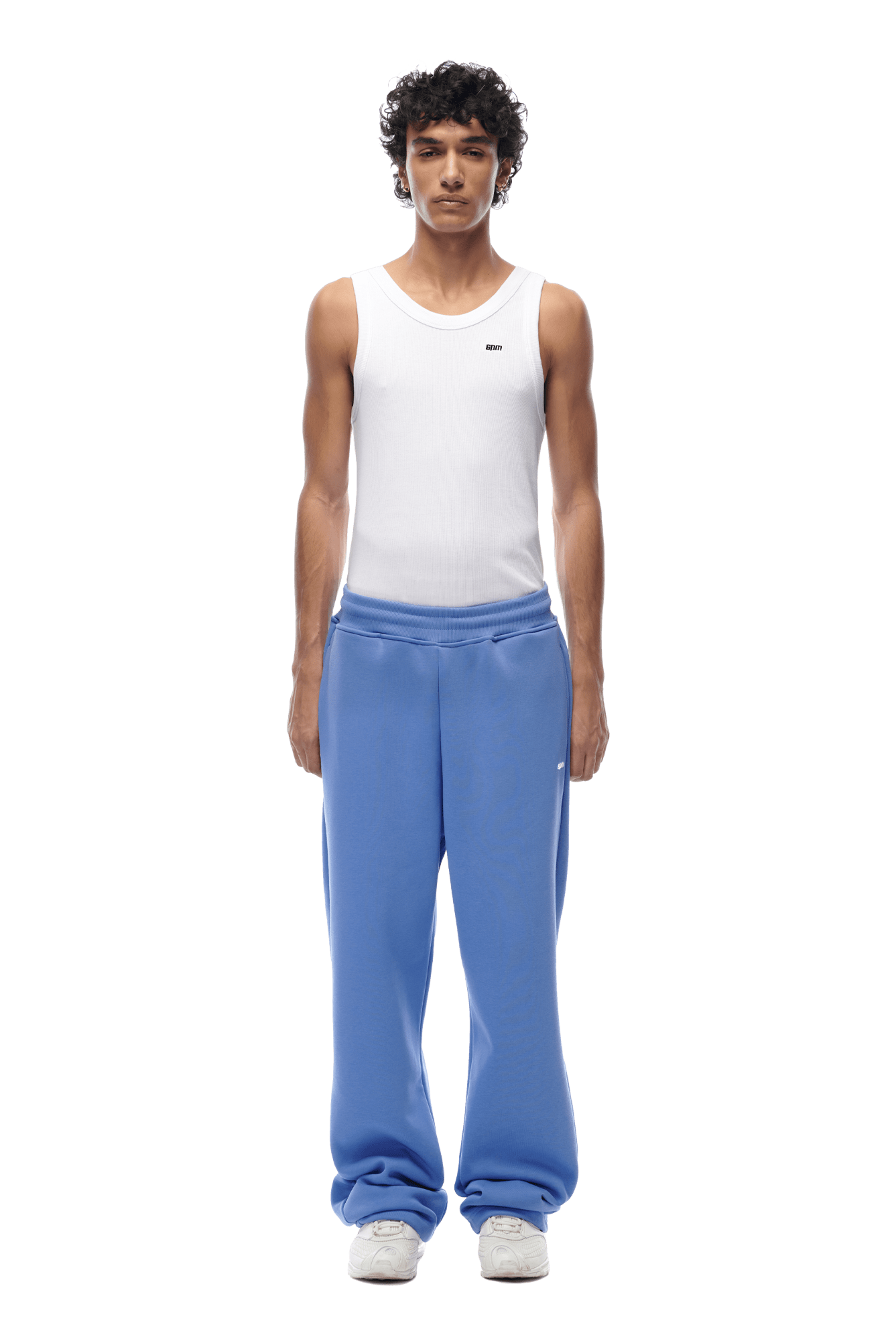 A man with short, curly hair stands facing forward, arms relaxed at his sides, wearing a white sleeveless tank top, 6PM OPEN LEG JOGGER PERSIAN BLUE sweatpants, and white sneakers. The background is transparent.