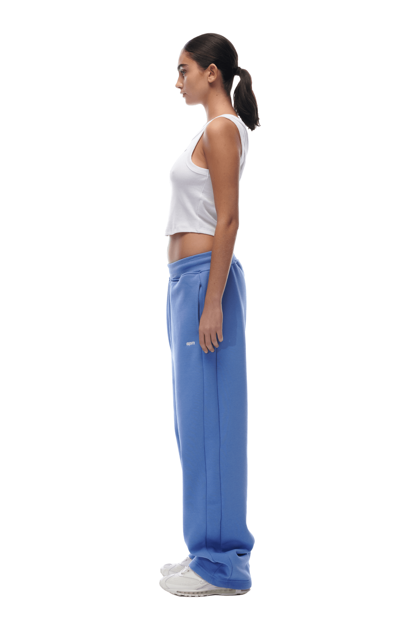 A woman in profile, facing left, wears a white sleeveless crop top and 6PM OPEN LEG JOGGER PERSIAN BLUE sweatpants with white sneakers. Her hair is tied in a low ponytail. The background is transparent.