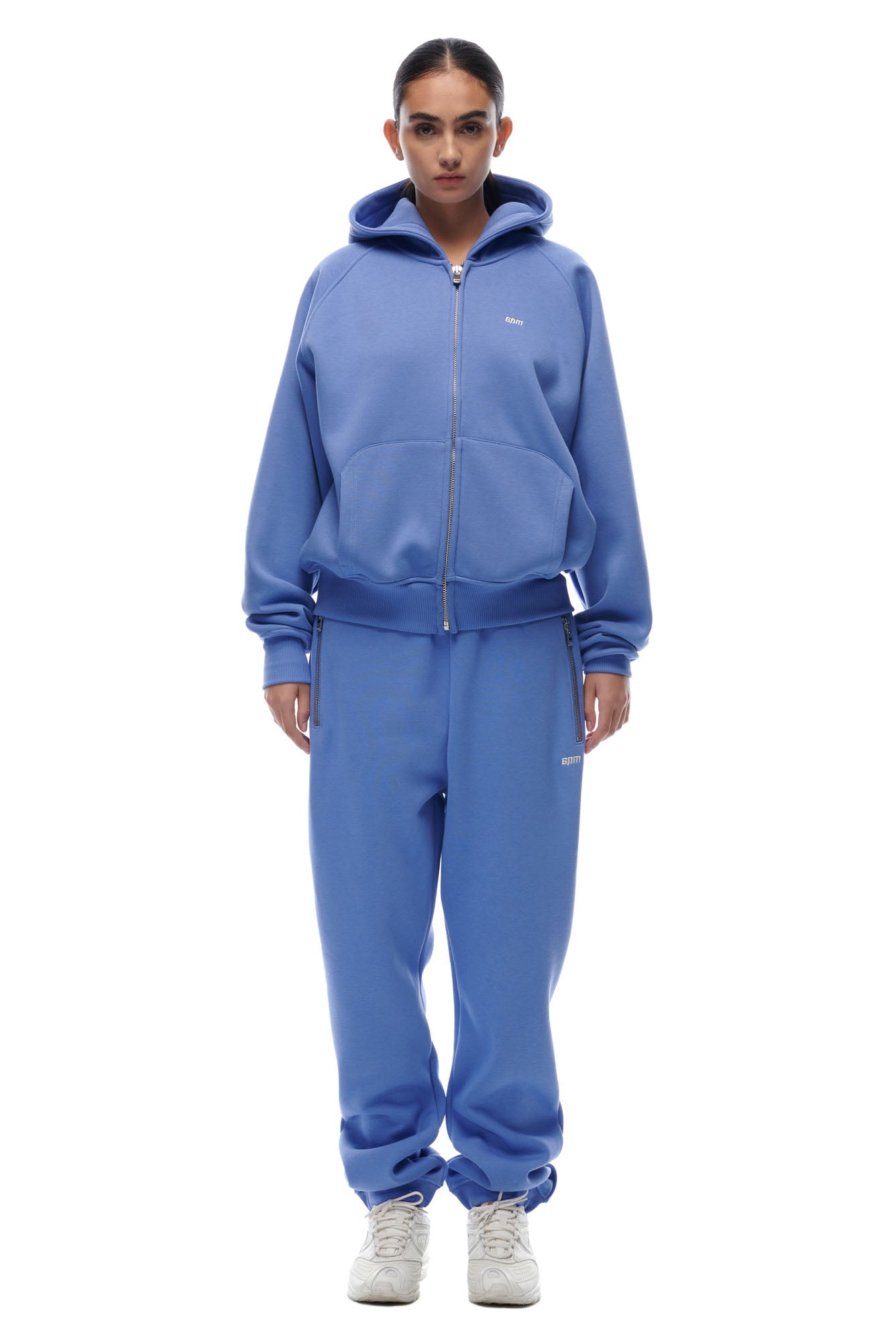 A woman faces forward against a white background, wearing the 6PM ZIP HOODIE PERSIAN BLUE and matching sweatpants with white sneakers. Her hair is tied back, highlighting the relaxed, casual fit—perfect for everyday wear.