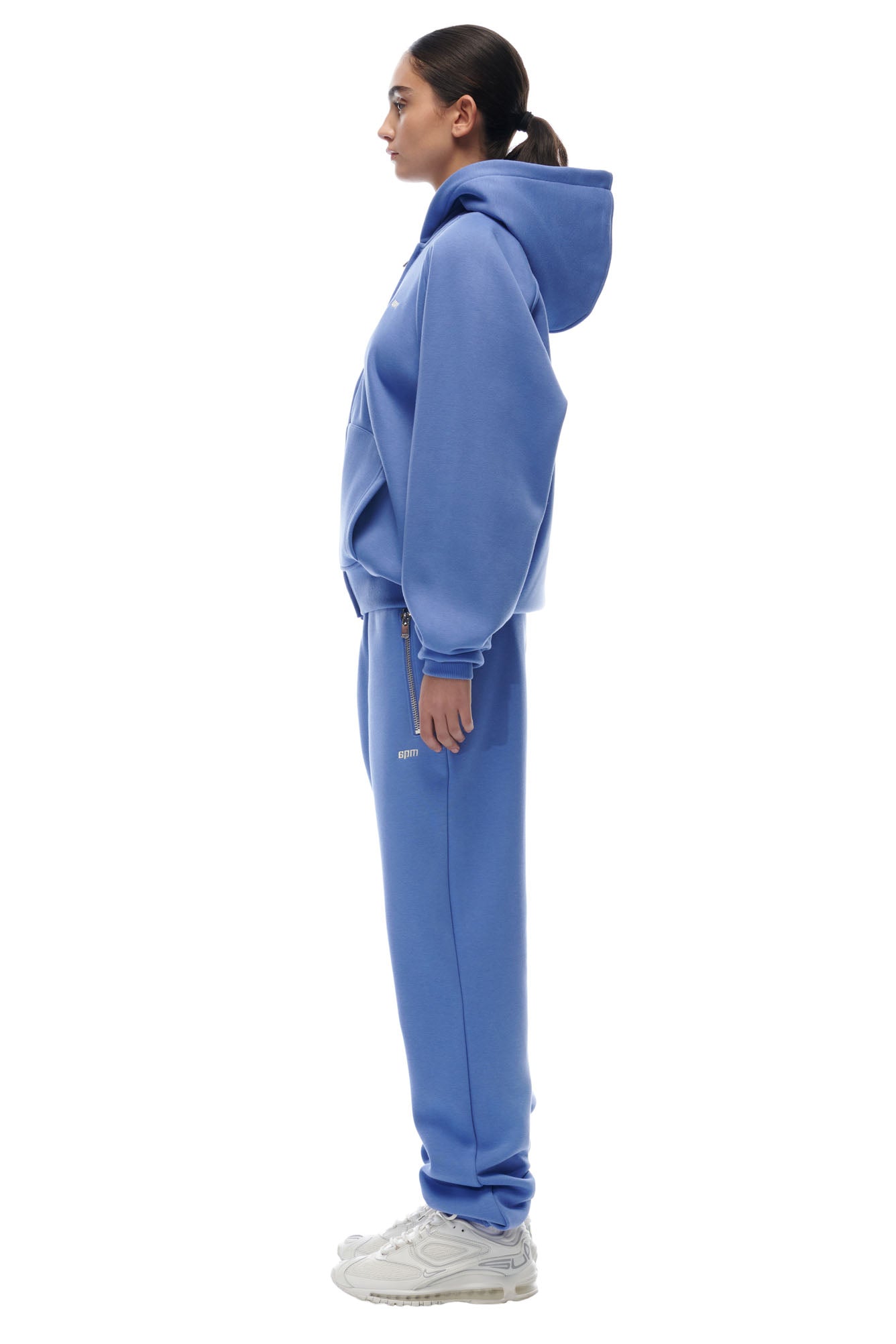 A woman in profile wears the 6PM ZIP HOODIE PERSIAN BLUE set with a clean finish and white sneakers. Her hair is in a low ponytail as she faces left against a plain white background.