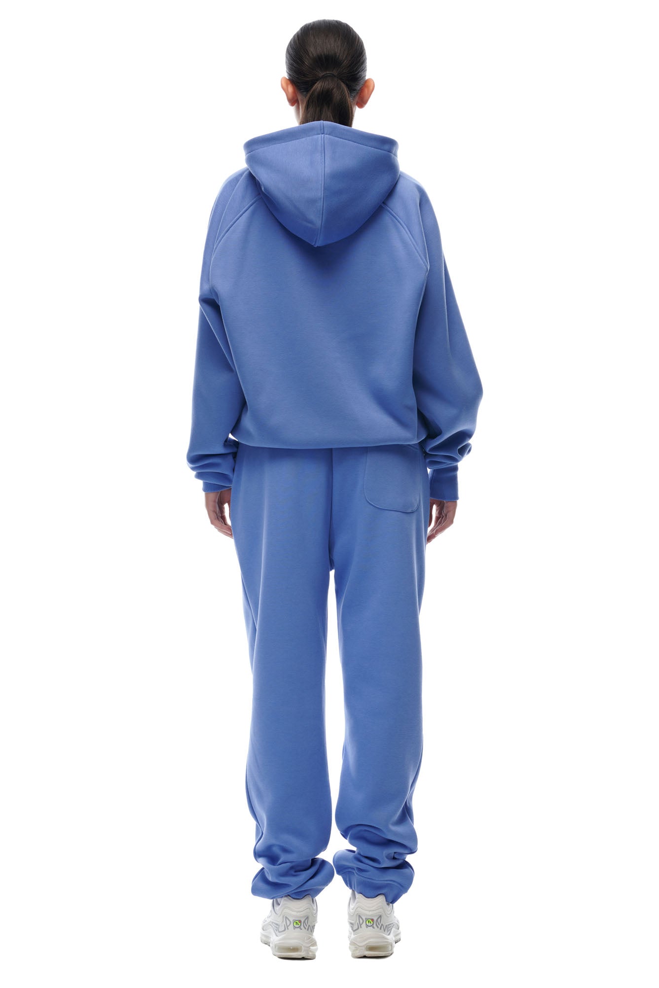 A person with long dark hair in a ponytail, wearing the 6PM ZIP HOODIE PERSIAN BLUE and matching sweatpants, stands facing away from the camera. They complete the look with white sneakers.