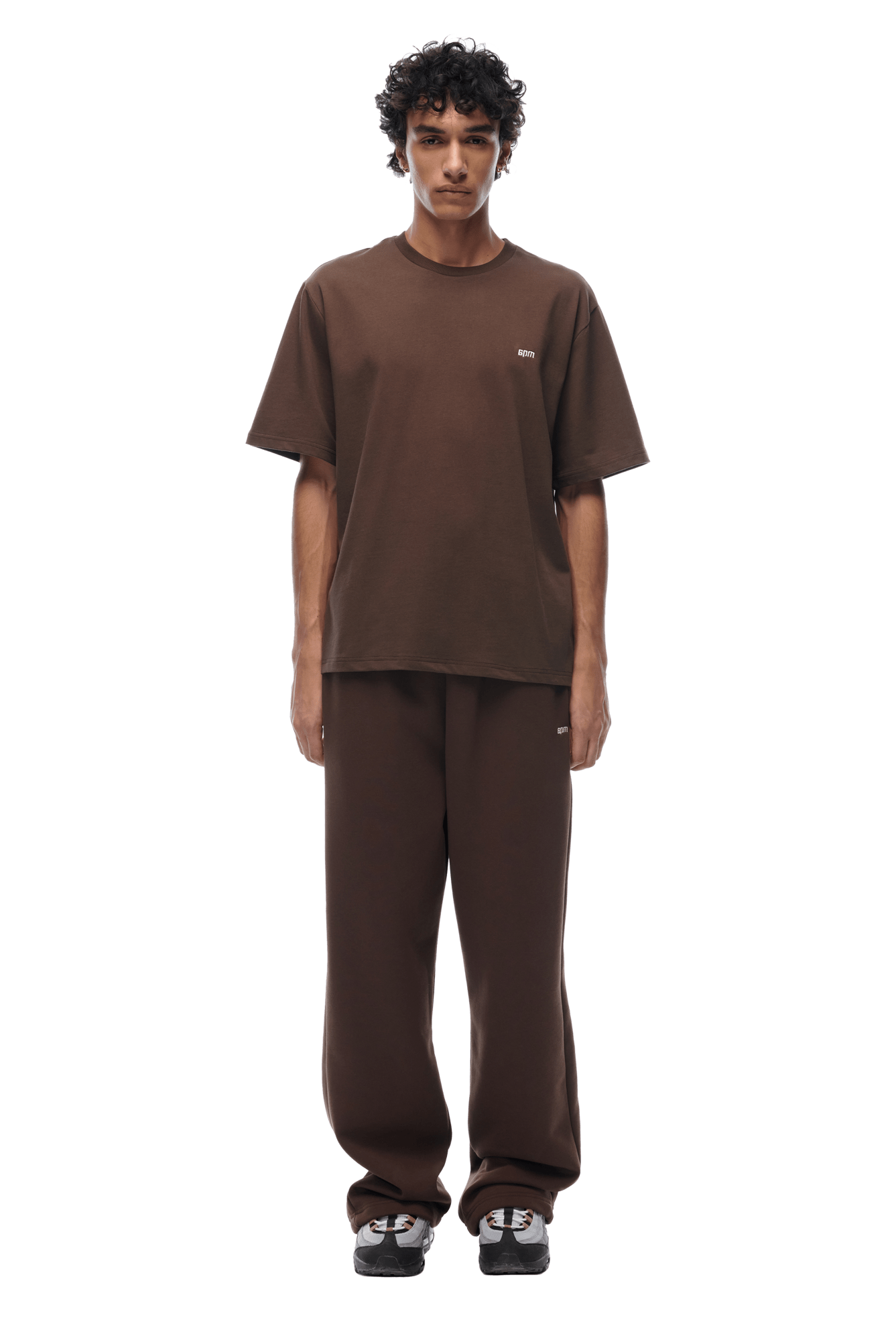 A young man with curly hair faces forward, wearing the 6PM T-SHIRT CHOCOLATE BROWN and matching brown sweatpants with black and white sneakers. The background is transparent.