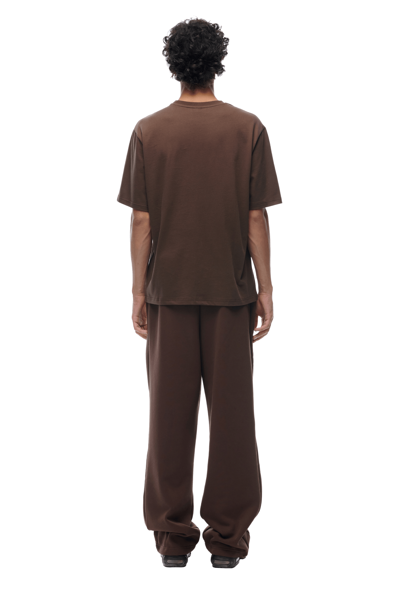 A person with short curly hair faces away, wearing a loose-fitting 6PM T-SHIRT CHOCOLATE BROWN and matching brown pants with black shoes, set against a plain background.