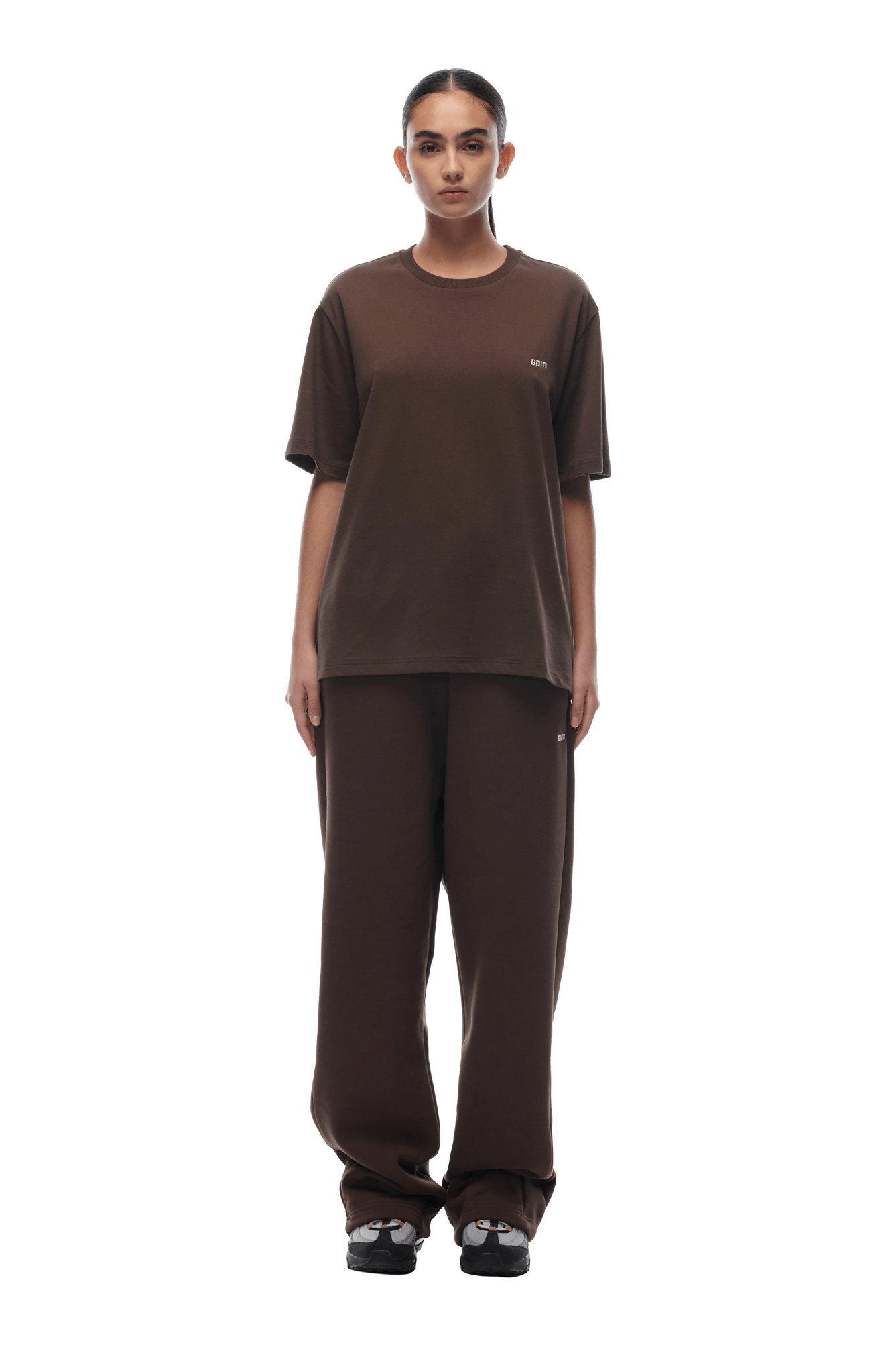 A person faces forward wearing the 6PM T-SHIRT CHOCOLATE BROWN with matching pants and black and white sneakers. The background is transparent.
