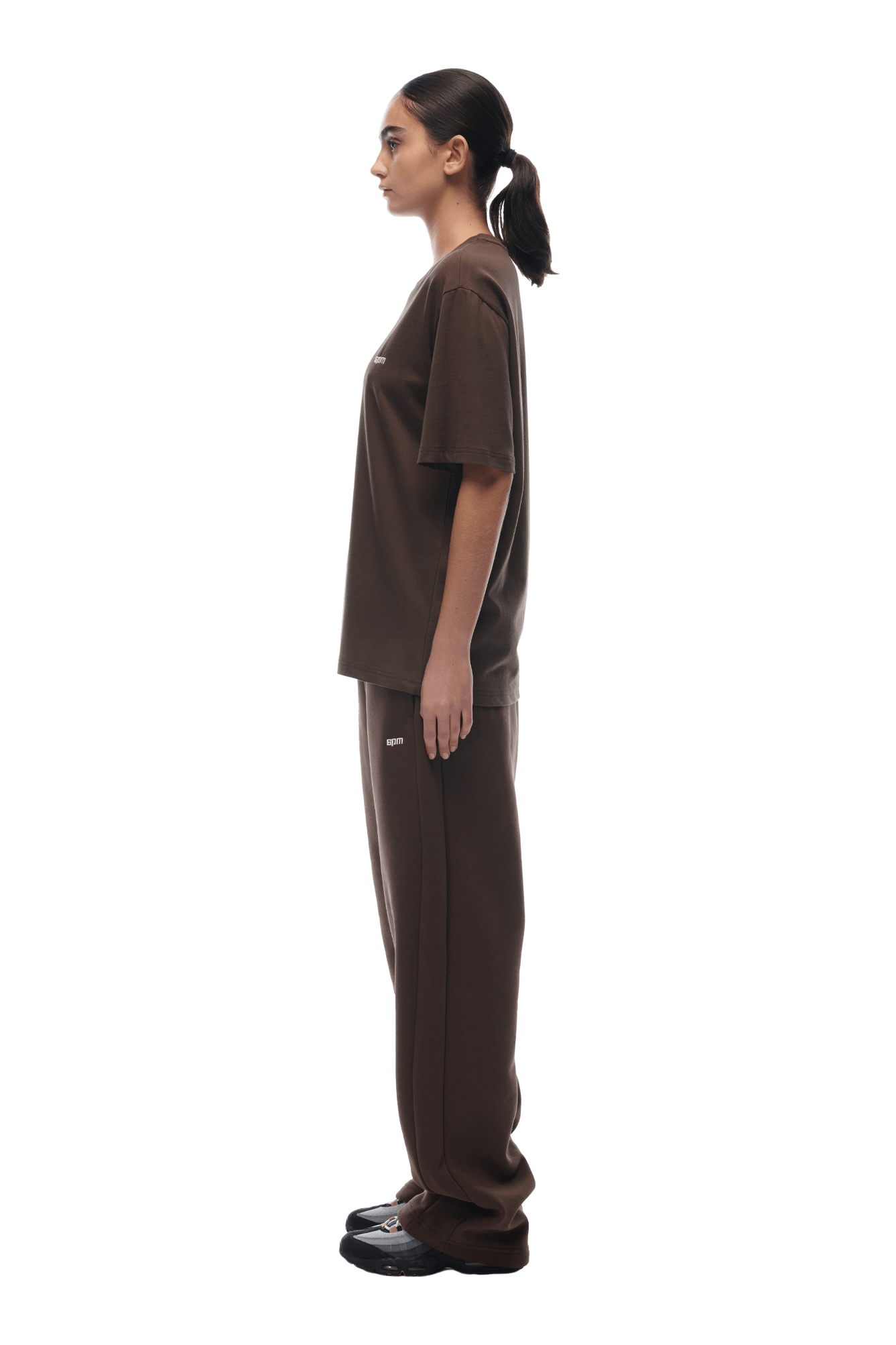 A woman in profile, facing left, wears the 6PM T-SHIRT CHOCOLATE BROWN with matching pants and black shoes. Her hair is tied back in a low ponytail. The background is transparent.