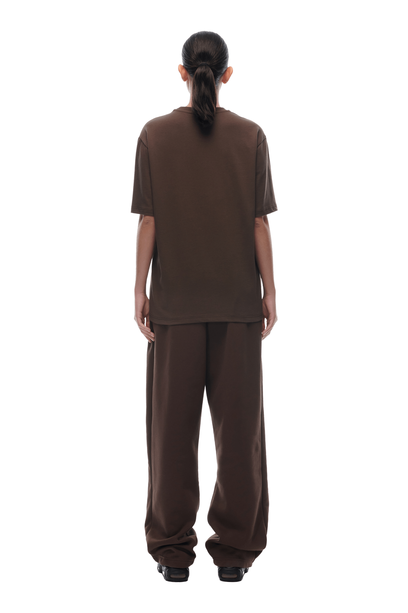 A person with long dark hair in a low ponytail faces away, wearing the oversized 6PM T-SHIRT CHOCOLATE BROWN and loose pants with black shoes, against a plain transparent background.