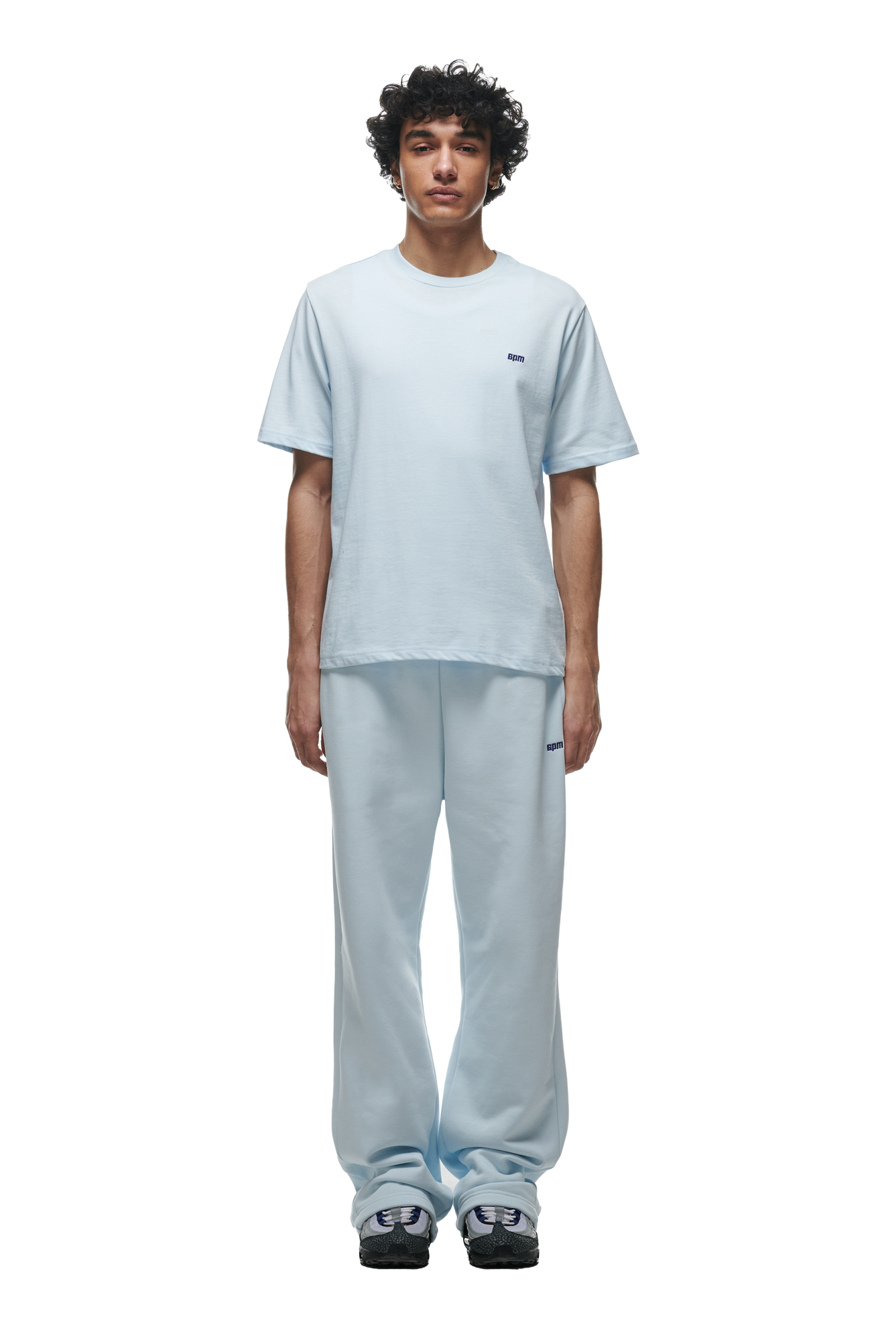 A person with curly hair stands facing forward, wearing a 6PM T-SHIRT ICE BLUE paired with matching loose sweatpants and black and white sneakers, set against a plain white background.