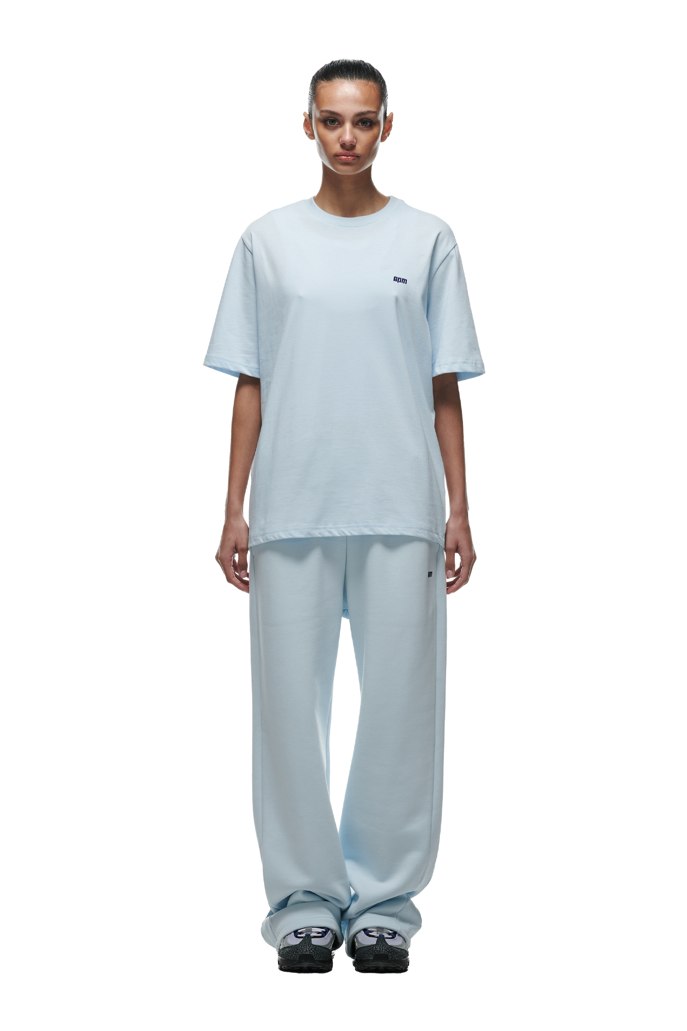 A woman stands facing forward against a plain white background, wearing the 6PM T-SHIRT ICE BLUE and matching wide-leg pants, paired with black and white sneakers.