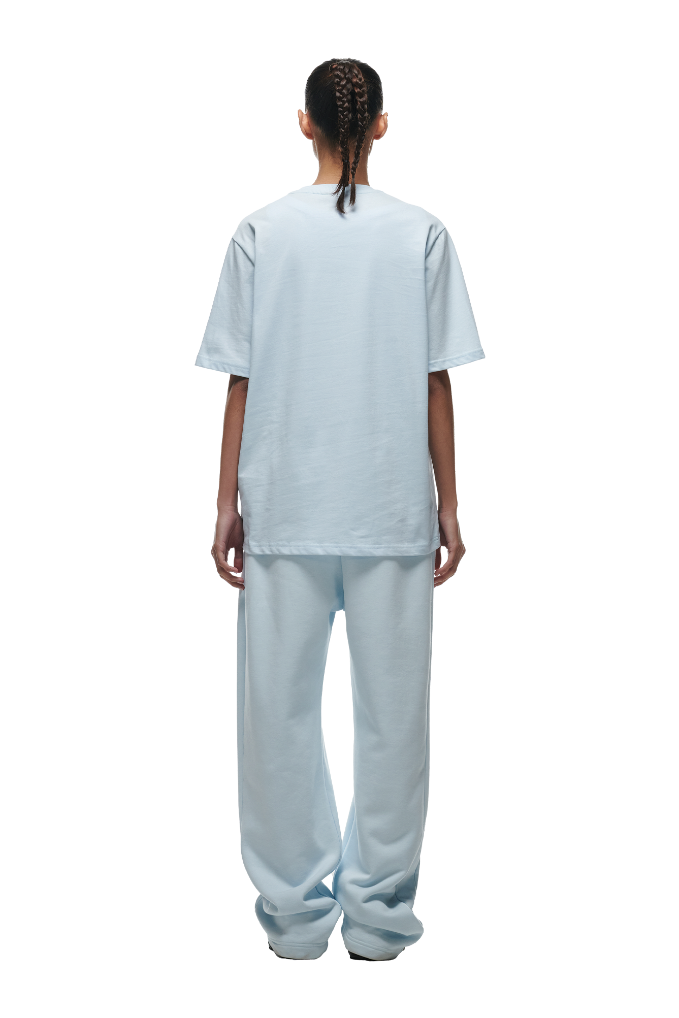 A person with braided hair faces away, wearing the 6PM T-SHIRT ICE BLUE and matching light blue pants with white sneakers against a plain background.