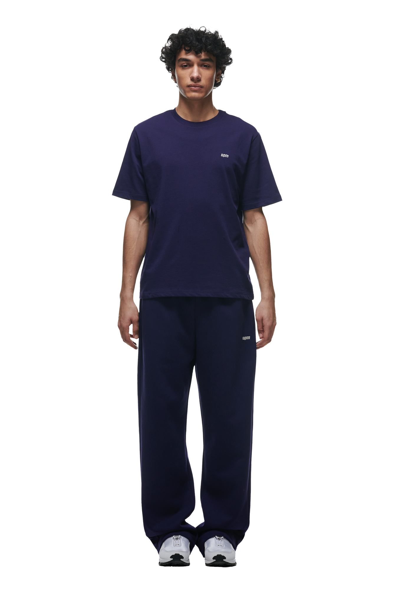 A person with curly hair stands facing forward, wearing the 6PM T-SHIRT ORBIT BLUE with matching navy pants and white sneakers, set against a plain white background.