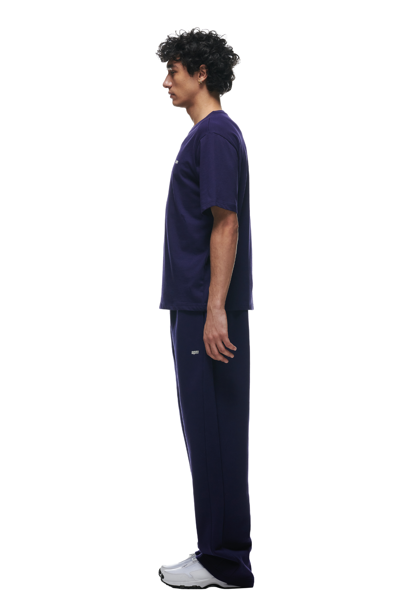 A man with curly hair stands in profile, wearing the 6PM T-SHIRT ORBIT BLUE and matching dark purple pants with white sneakers against a white background.