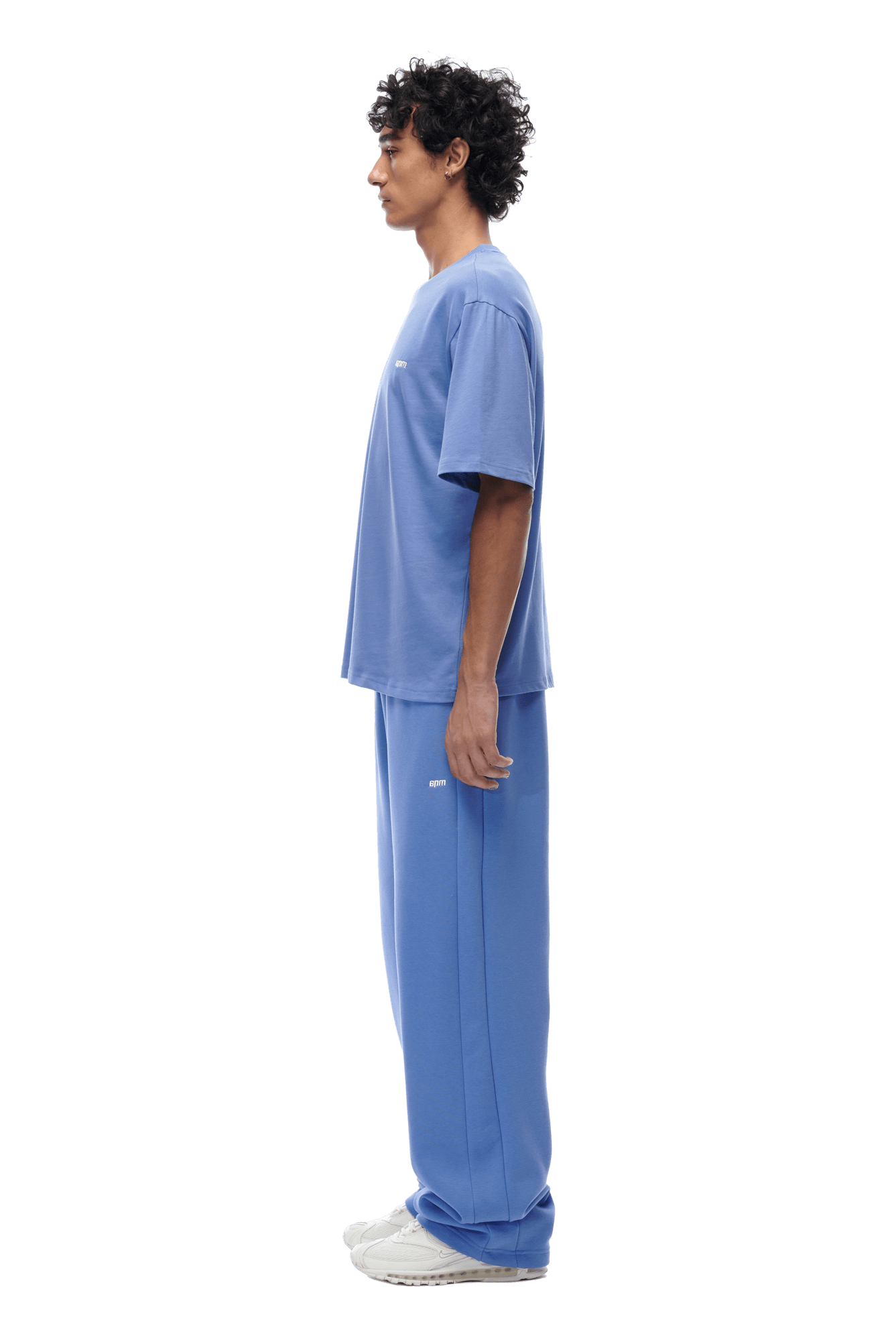 A curly-haired man stands in profile facing left, wearing the 6PM T-SHIRT PERSIAN BLUE with matching blue sweatpants and white sneakers. The background is transparent.