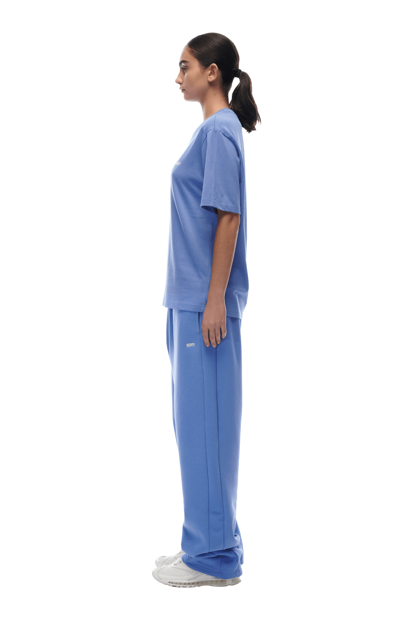 A person stands in profile, wearing the 6PM T-SHIRT PERSIAN BLUE and matching light blue sweatpants with white sneakers against a plain background.