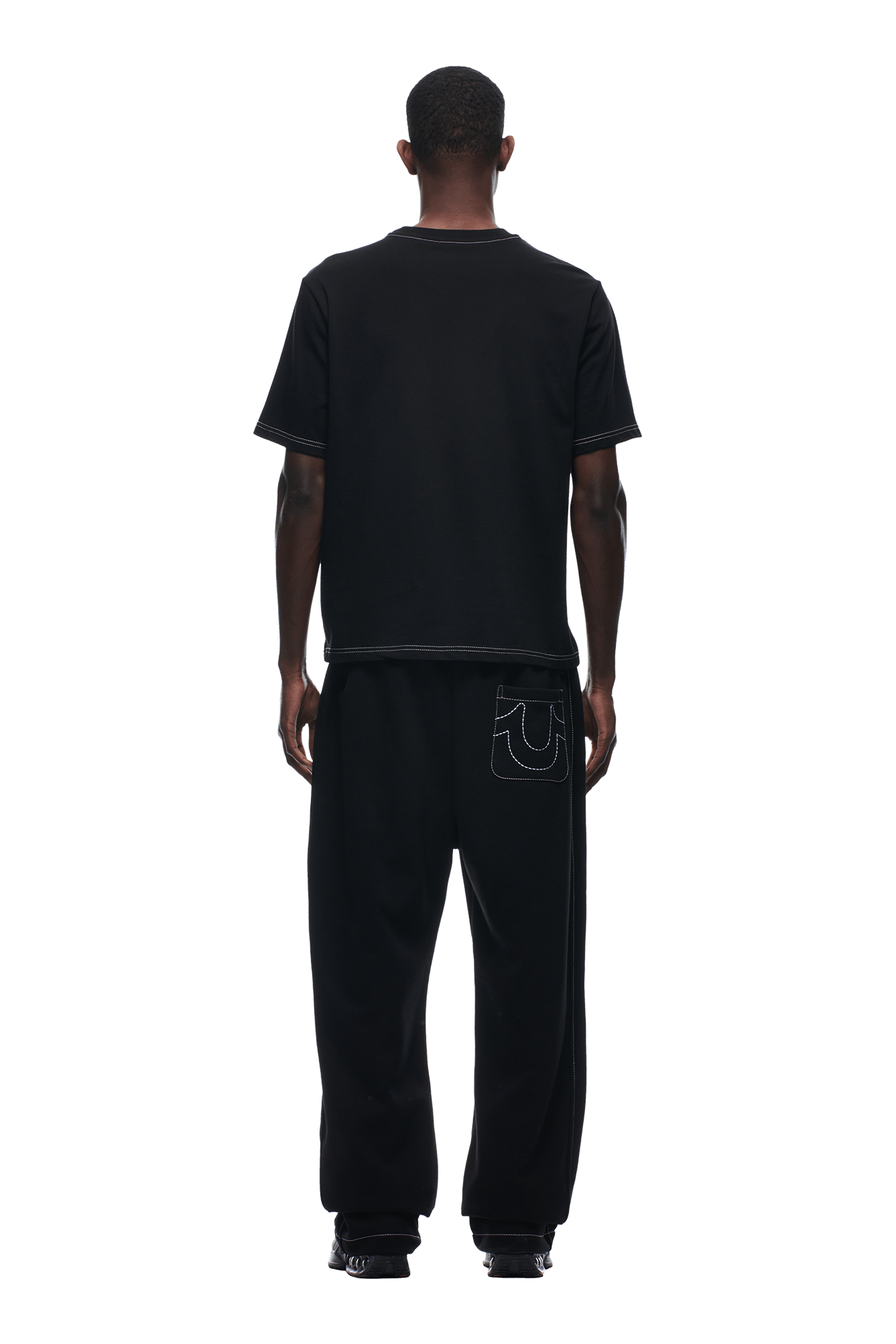 A person faces away, wearing the 6PM BUDDHA T-SHIRT BLACK and loose black pants with a large stitched design on the back pocket, against a plain white background.