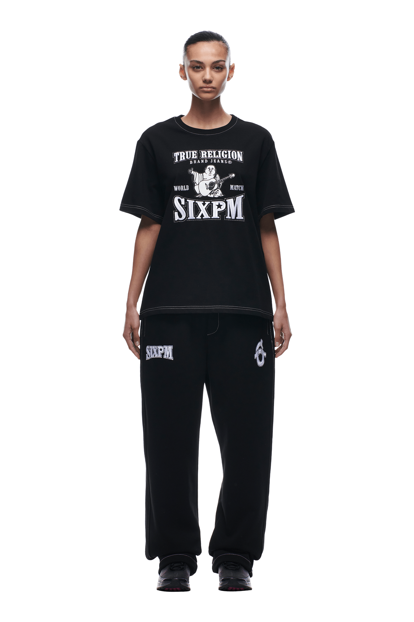 A woman faces forward wearing the BUDDHA T-SHIRT BLACK by 6PM, paired with matching black sweatpants featuring 6PM patches. She completes the look with slicked-back hair and black sneakers.