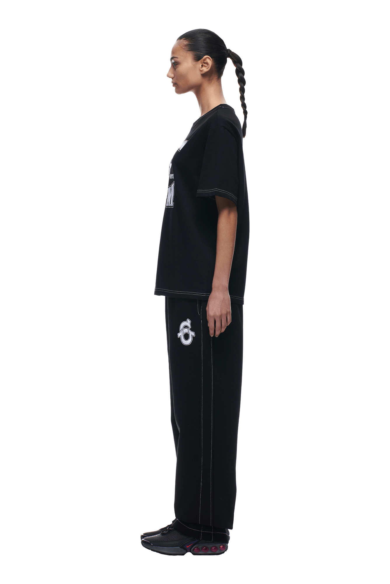 A person with a braided ponytail stands in profile, wearing the 6PM BUDDHA T-SHIRT BLACK, wide black pants with a white logo on the thigh, and black sneakers, against a plain white background.