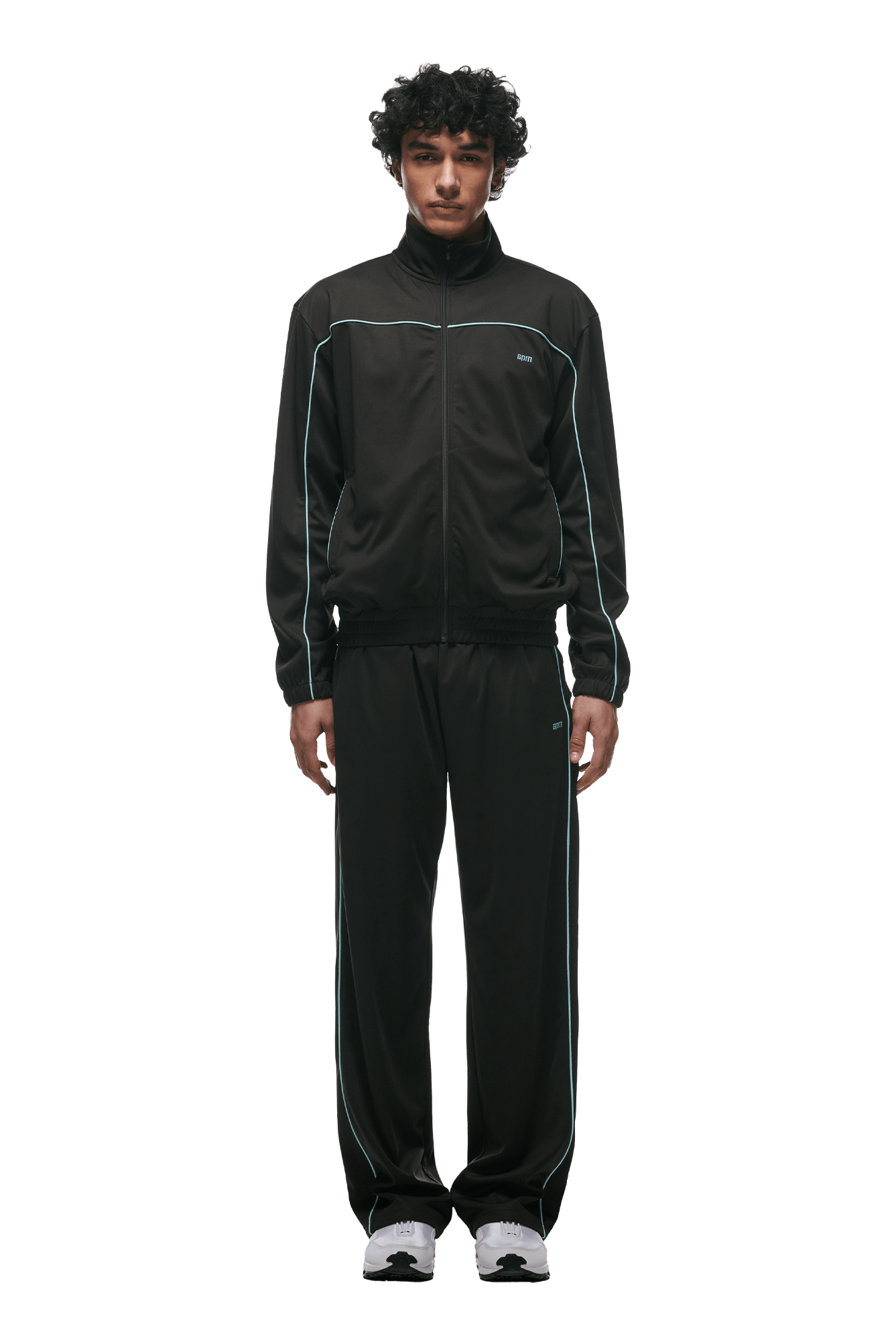 A person stands facing forward in the 6PM+ TRAINER JACKET ANTHRACITE by 6PM, featuring white piping and zipped pockets, paired with white athletic shoes—set against a plain green background for a sleek, urban look.